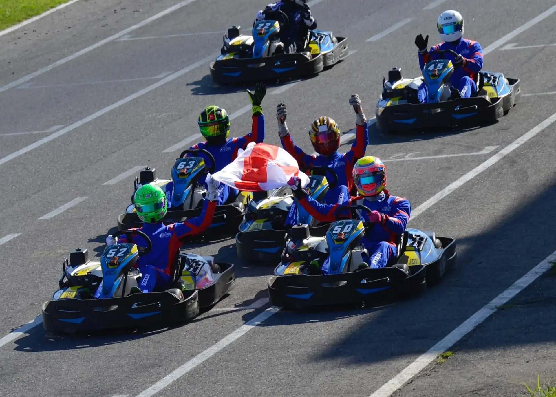 A group of go-kart drivers in racing suits and helmets drive on a track, with the front driver holding up a flag and several drivers raising their hands in celebration.