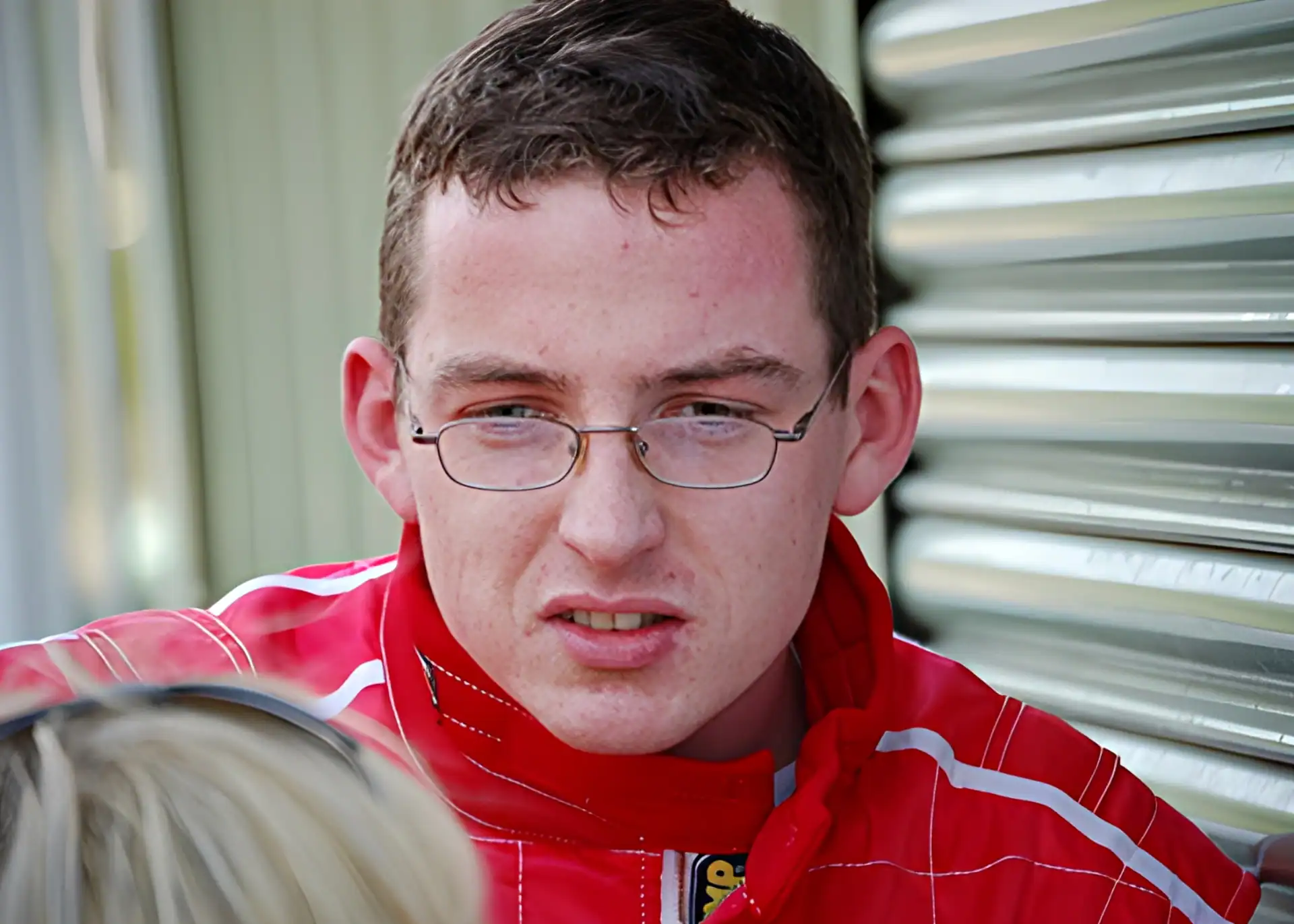 A man wearing glasses and a red racing suit stands near a corrugated metal wall, looking slightly to the side.