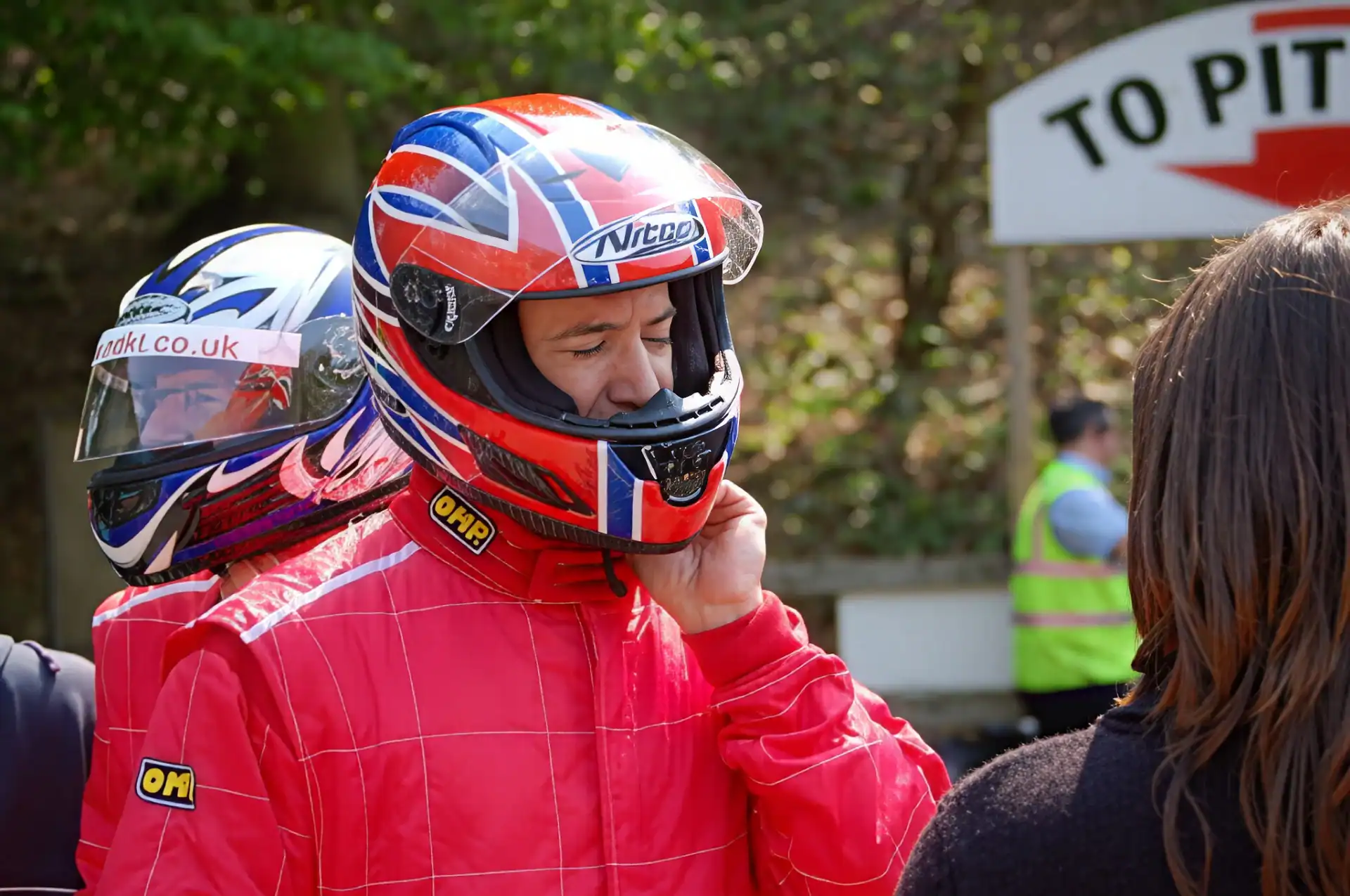 A person in a red racing suit and helmet adjusts their chin strap, standing outdoors near others, with a "To Pits" sign visible in the background.