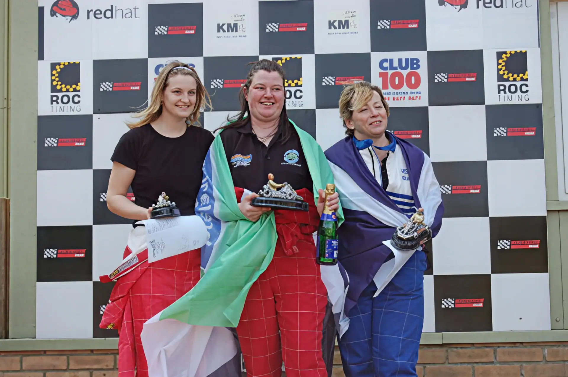 Three women stand on a winners' rostrum holding trophies and wrapped in flags, with a backdrop of sponsor logos behind them.