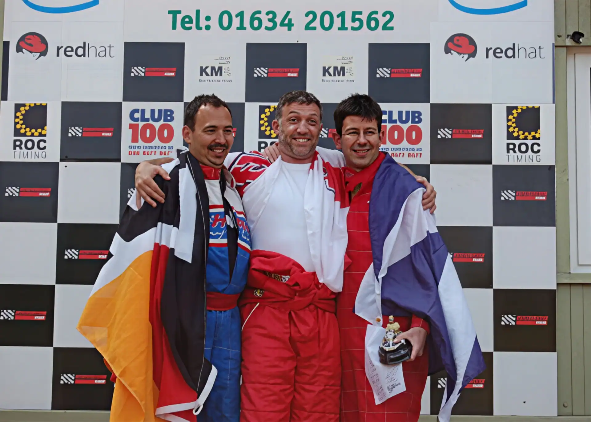 Three men in racing suits stand smiling on a podium, each holding a flag, with a chequered backdrop displaying sponsor logos and a phone number behind them.