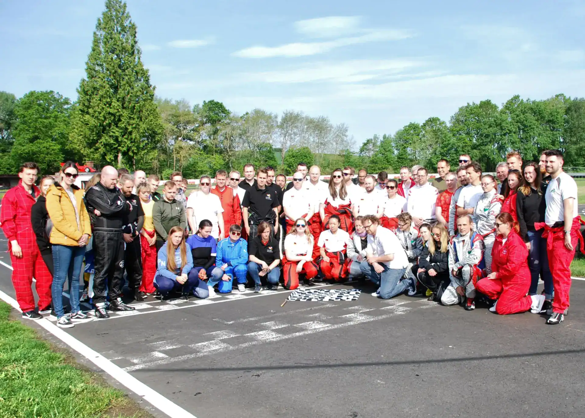 A large group of people, many in racing suits, pose together on a racetrack near a chequered finish line, with trees and blue sky in the background.