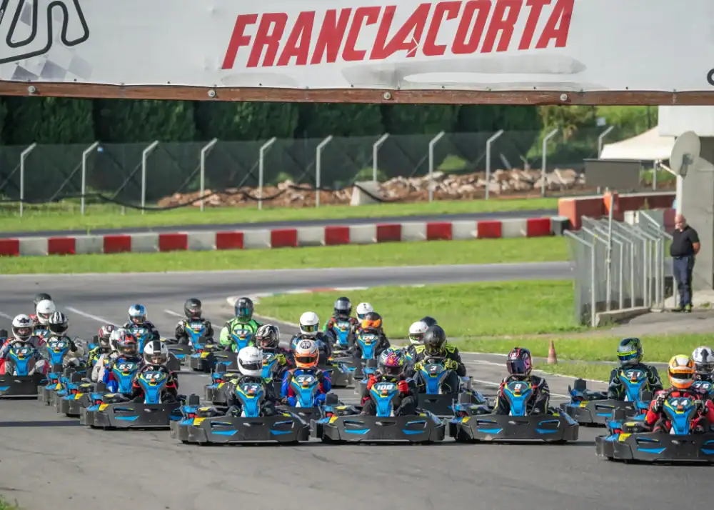 A group of go-kart racers wearing helmets and suits start a race on an outdoor track under a sign reading "FRANCIACORTA".