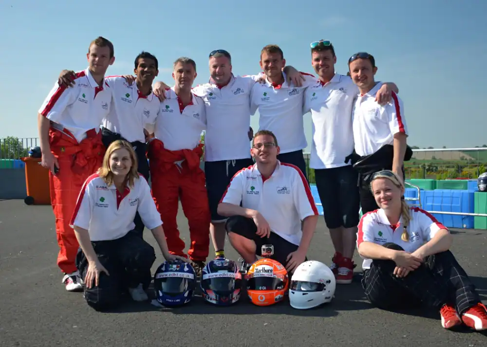 A group of nine people in matching team shirts pose outdoors on tarmac with four racing helmets placed in front of them.