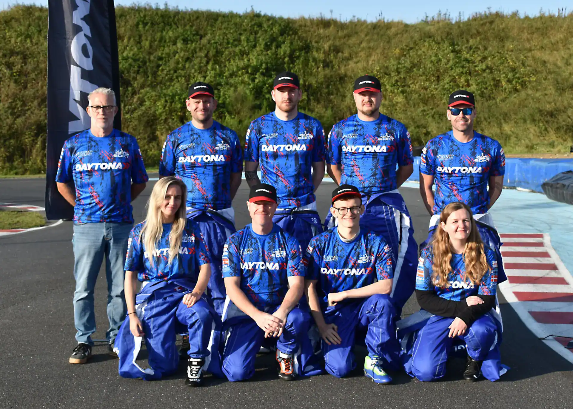 Nine people in matching blue Daytona racing uniforms pose for a group photo outdoors on a go-kart track, with greenery in the background.