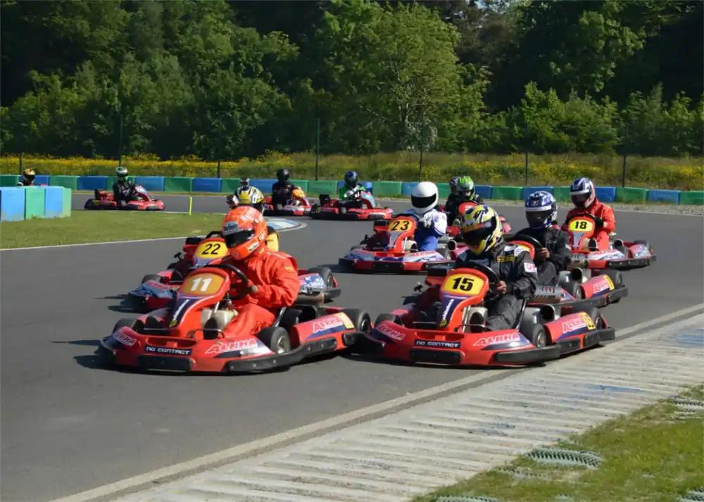 A group of go-kart racers in helmets and suits drive on a track surrounded by greenery, with numbered karts visible in the foreground.