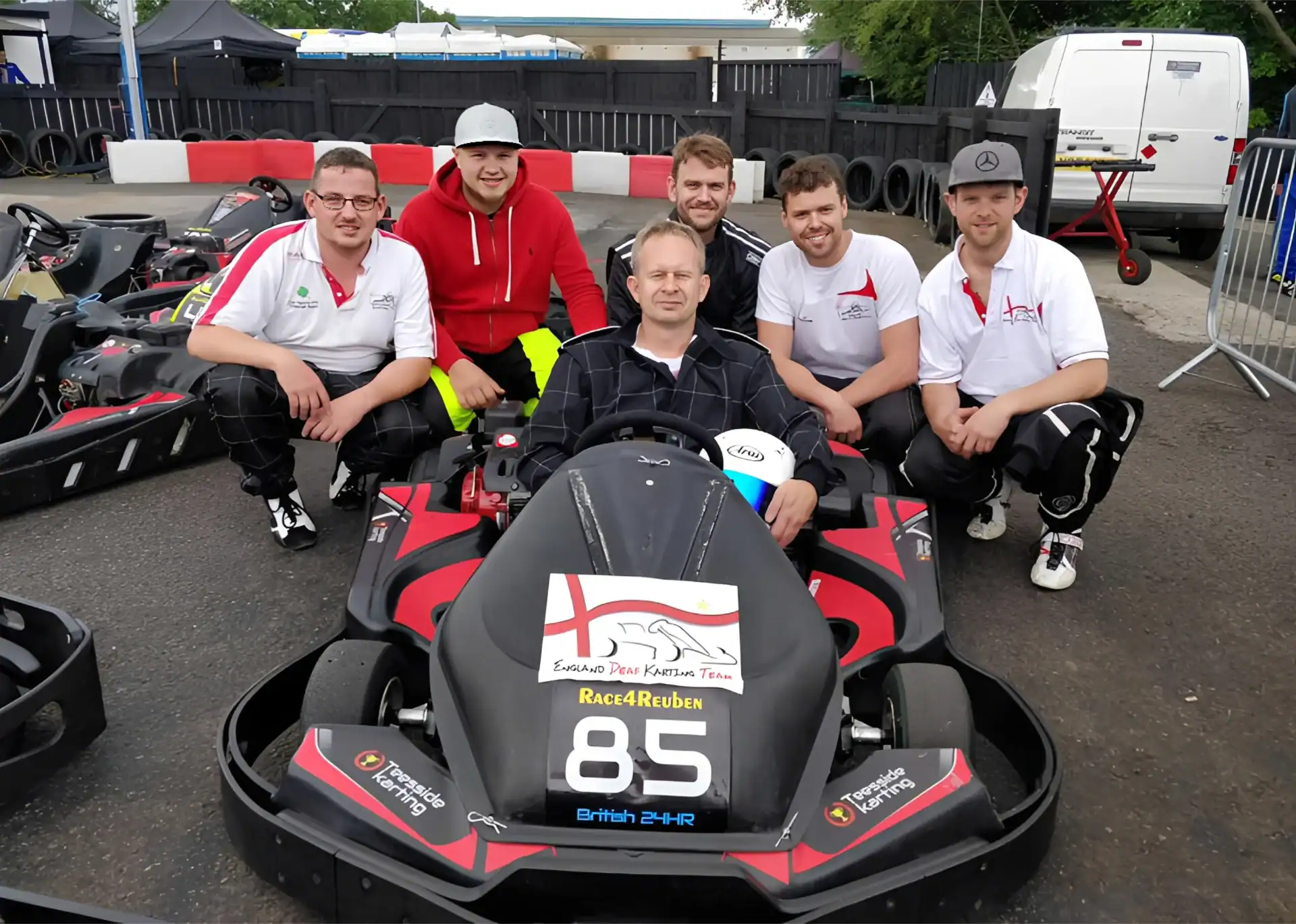 Six men pose around a go-kart labelled “85” at a karting track; one man sits in the kart while the others kneel or stand behind him, all dressed in casual sportswear.