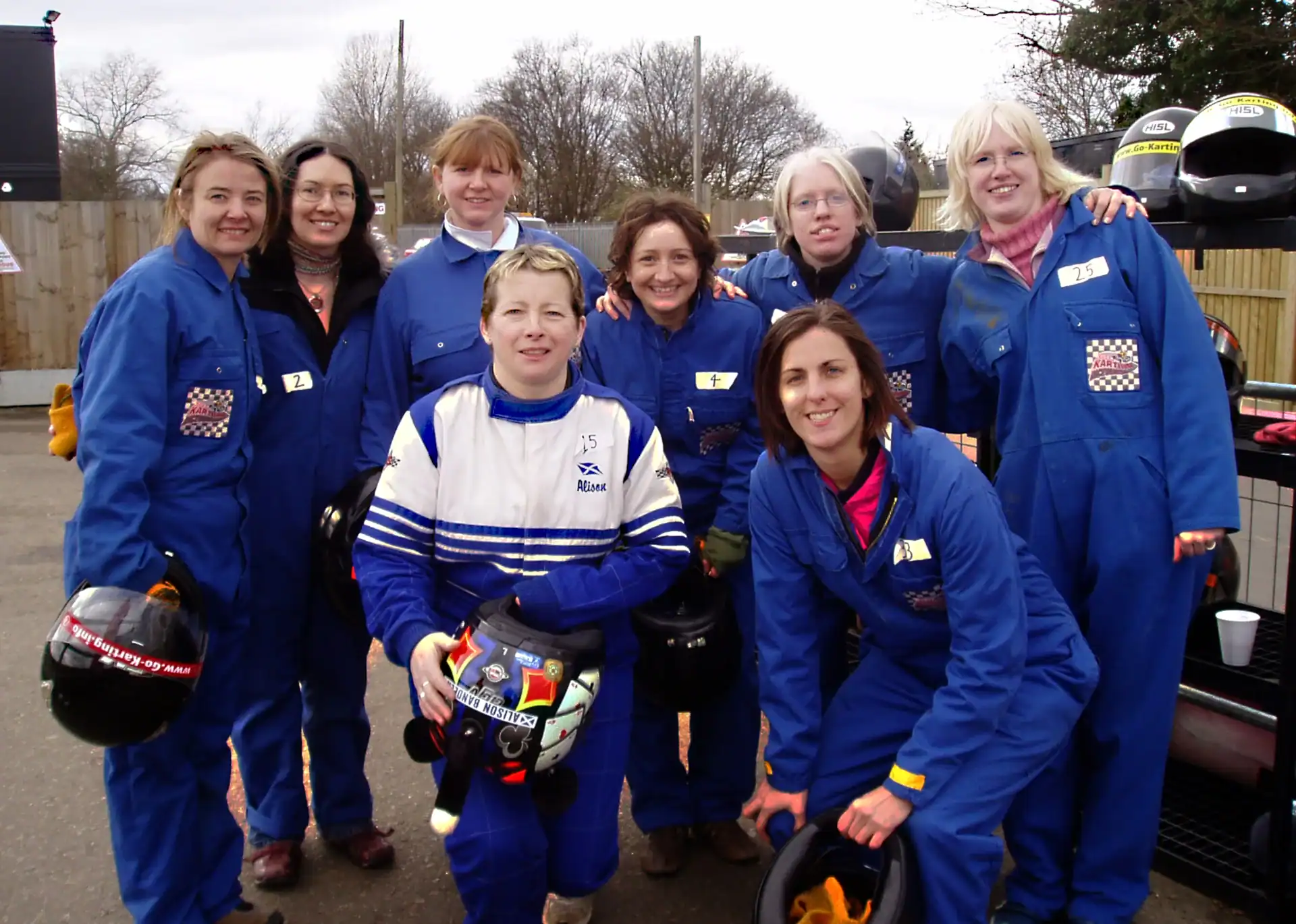 Eight women wearing blue racing suits and holding helmets pose together outdoors, with trees and helmets in the background.