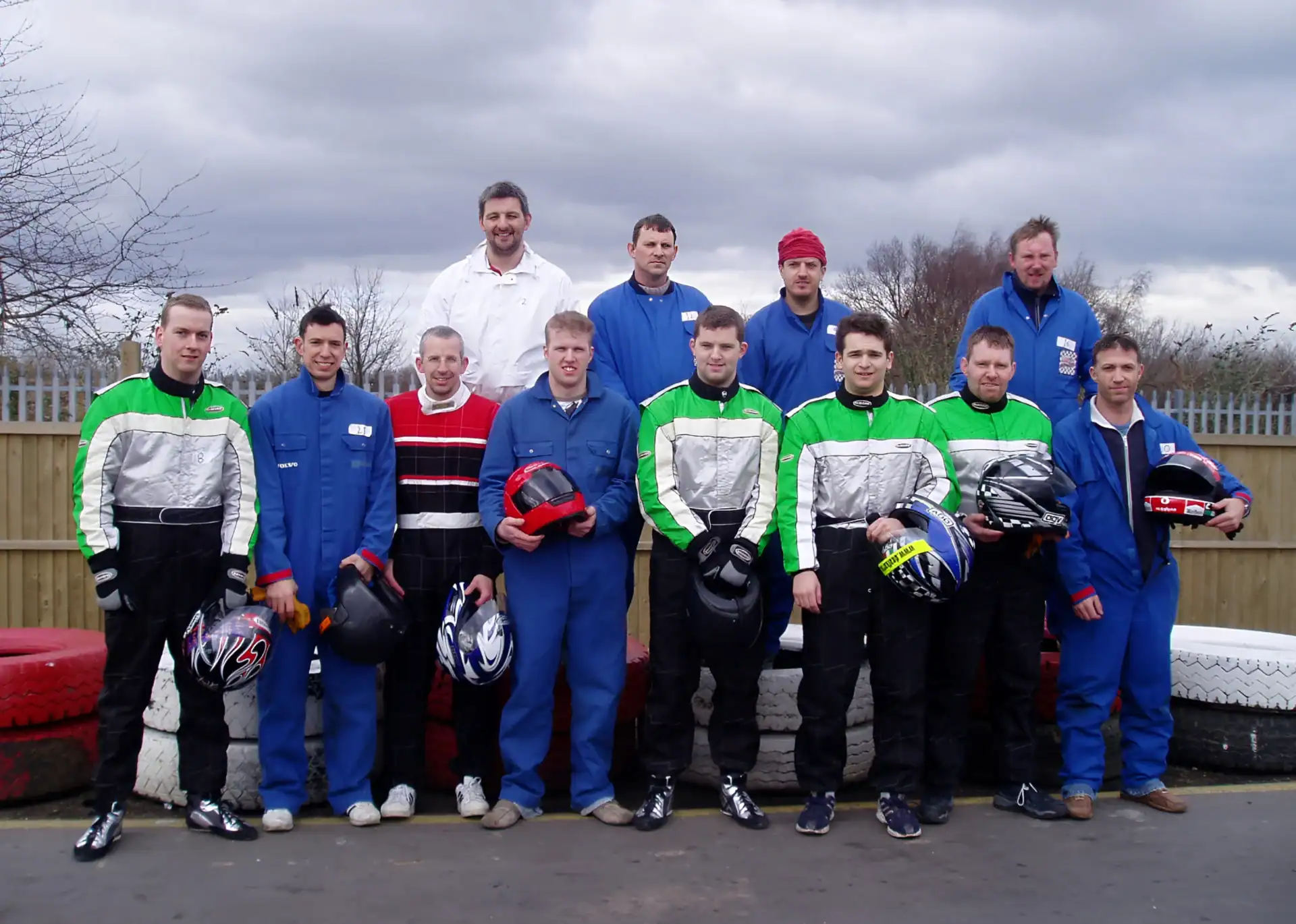 A group of thirteen men, some in blue or green racing suits and others in casual clothes, pose outdoors holding motorbike helmets near stacked tyres.