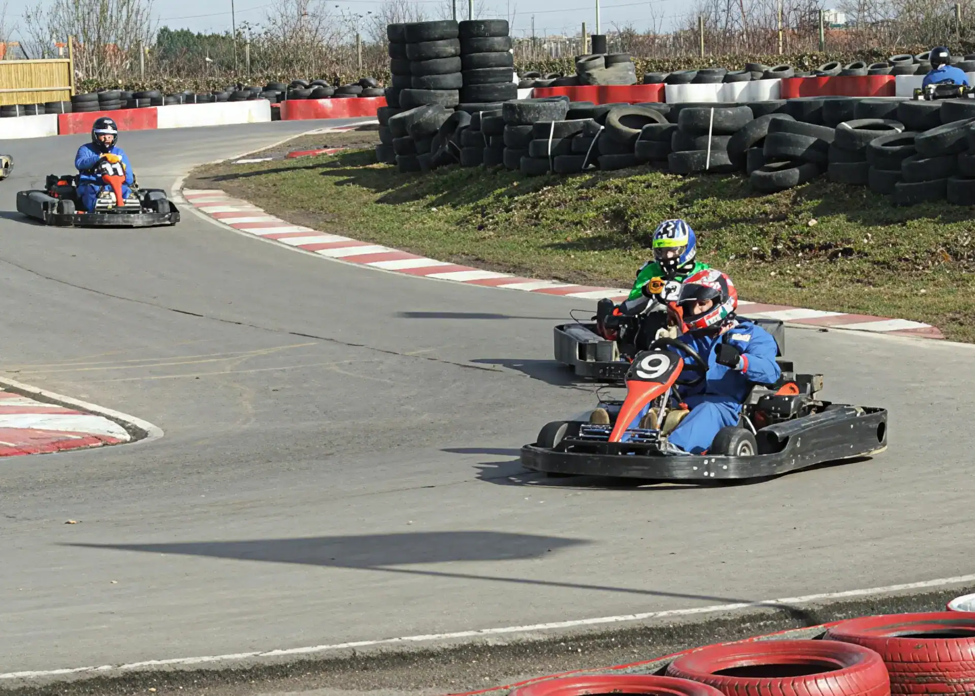 Three people racing go-karts on an outdoor track, with protective helmets and tyres lining the course. The leading kart is marked with the number 9.