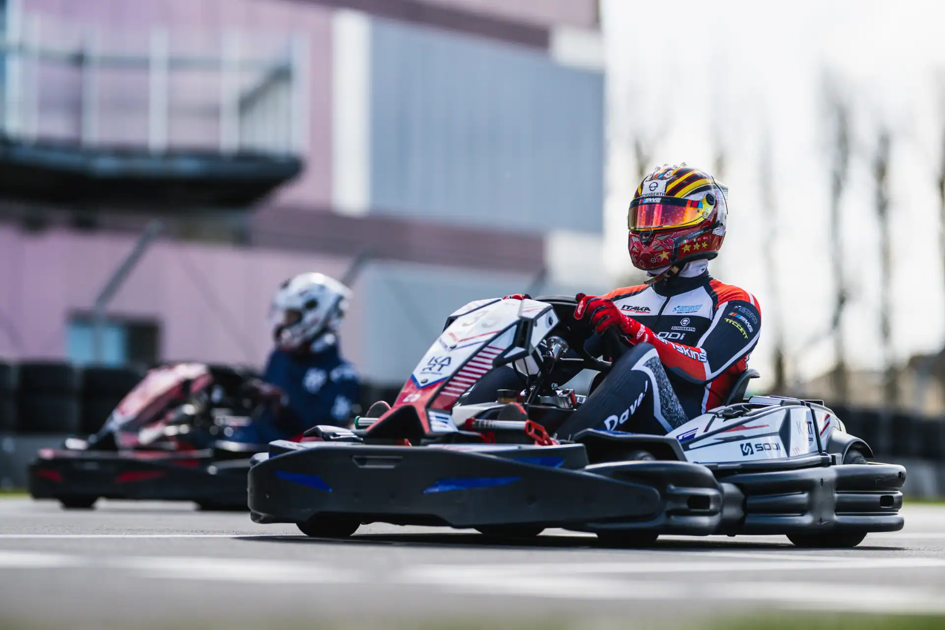 Two go-kart drivers wearing helmets and racing suits compete on an outdoor track, with one driver in the foreground and another in the background.
