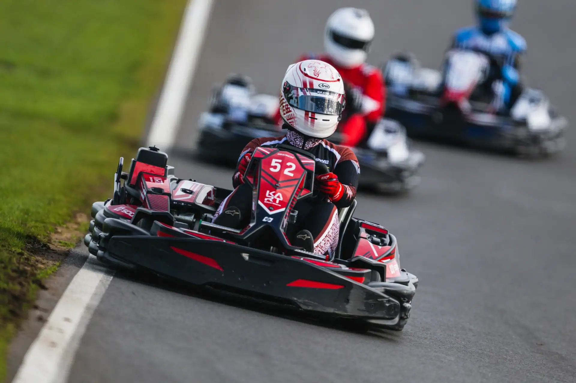 Person in go-kart number 52 driving on a racetrack, followed by other racers in go-karts, with grass visible to the side.
