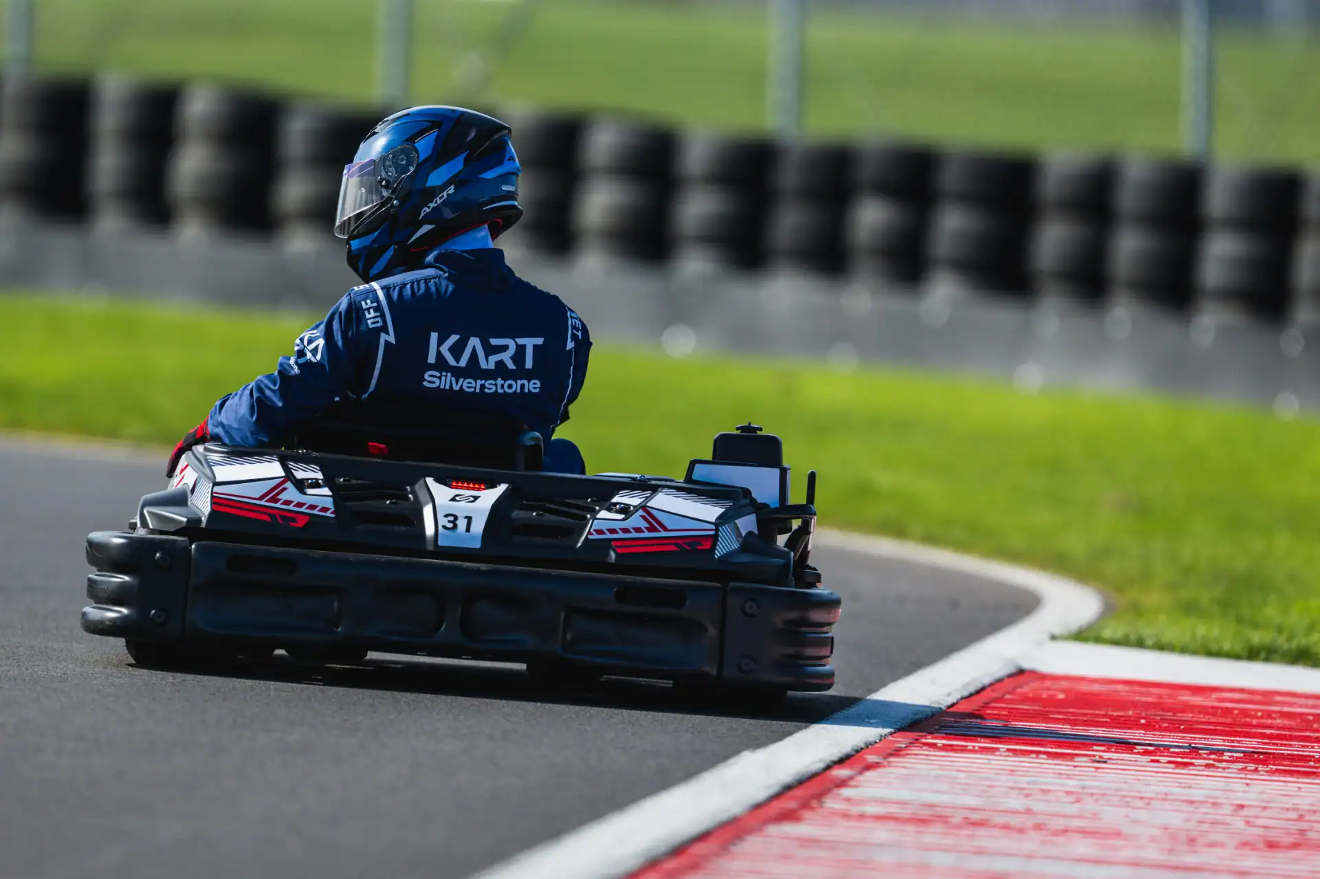 Person driving a go-kart on a racetrack, wearing a blue racing suit and helmet, with the word "KART Silverstone" visible on their back.