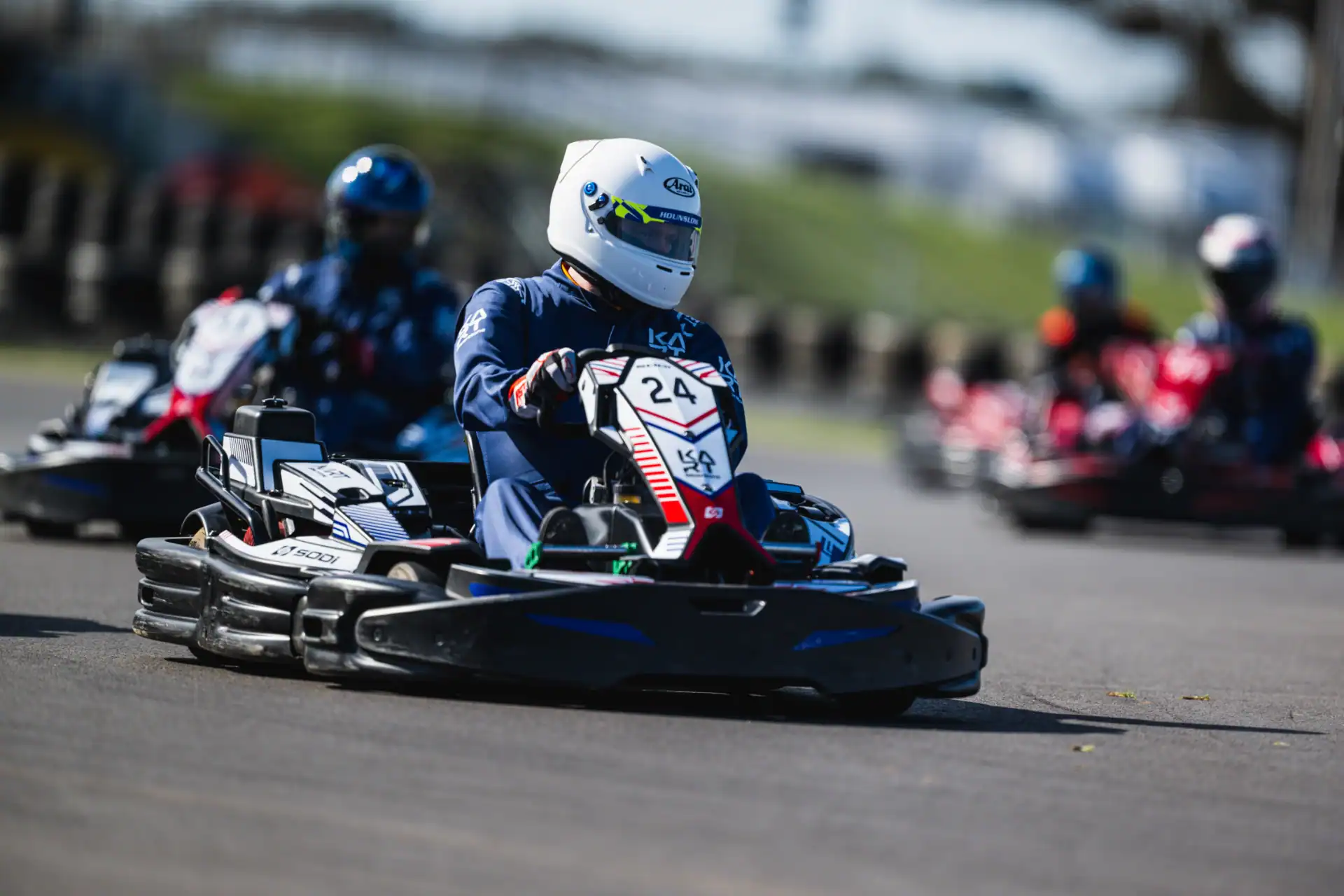 Person driving a go-kart labelled 24 on an outdoor track, wearing a white helmet and blue racing suit, with other racers in the background.