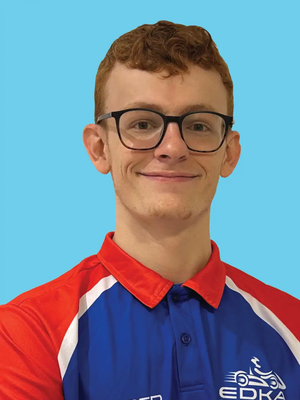 A person with short red hair and glasses wearing a blue, red, and white EDKA polo shirt stands in front of a solid light blue background.