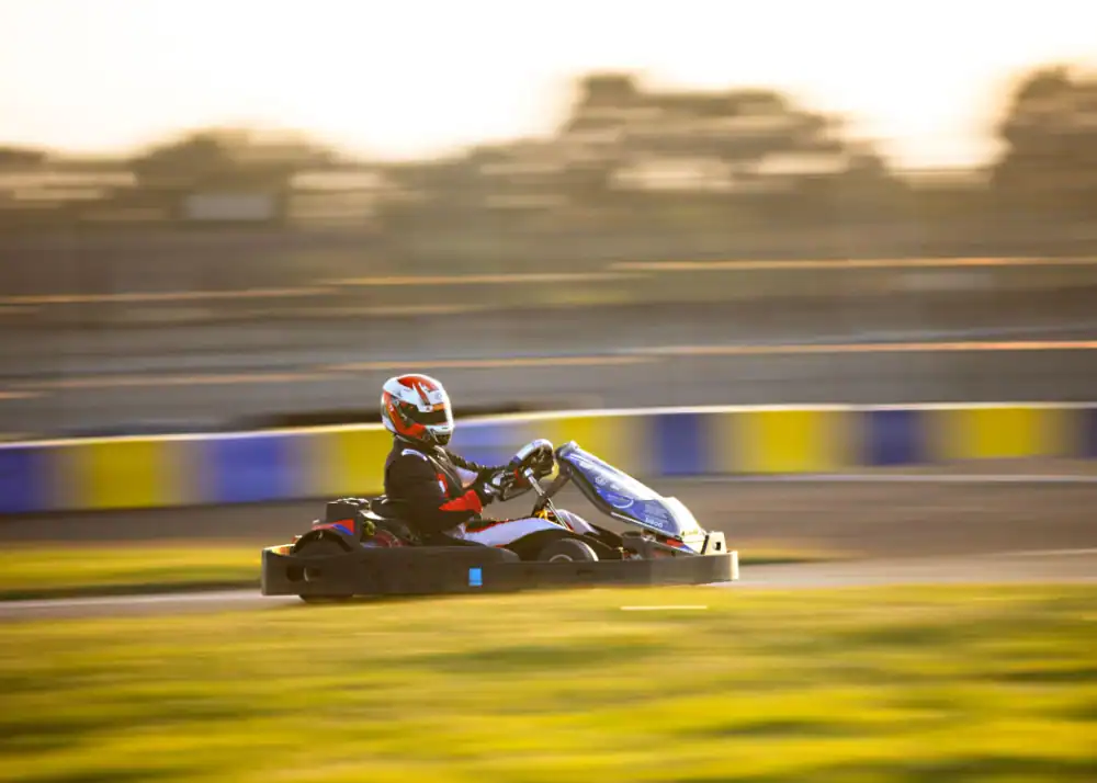 A person wearing a helmet and racing suit drives a go-kart at high speed on an outdoor track, with blurred background and sunlight.