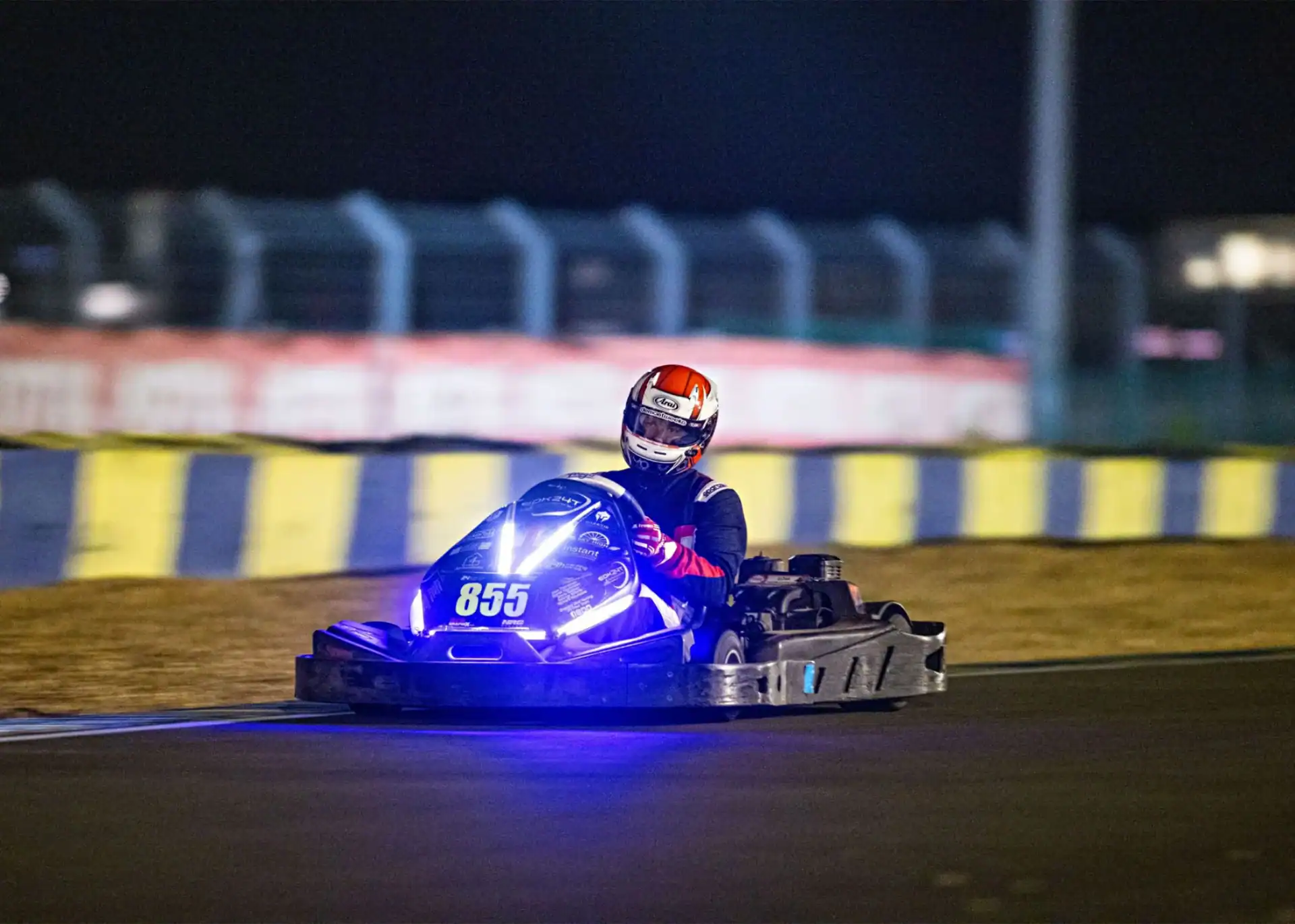 A person wearing a red and white helmet drives a go-kart with blue lights and the number 855 on a racetrack at night.