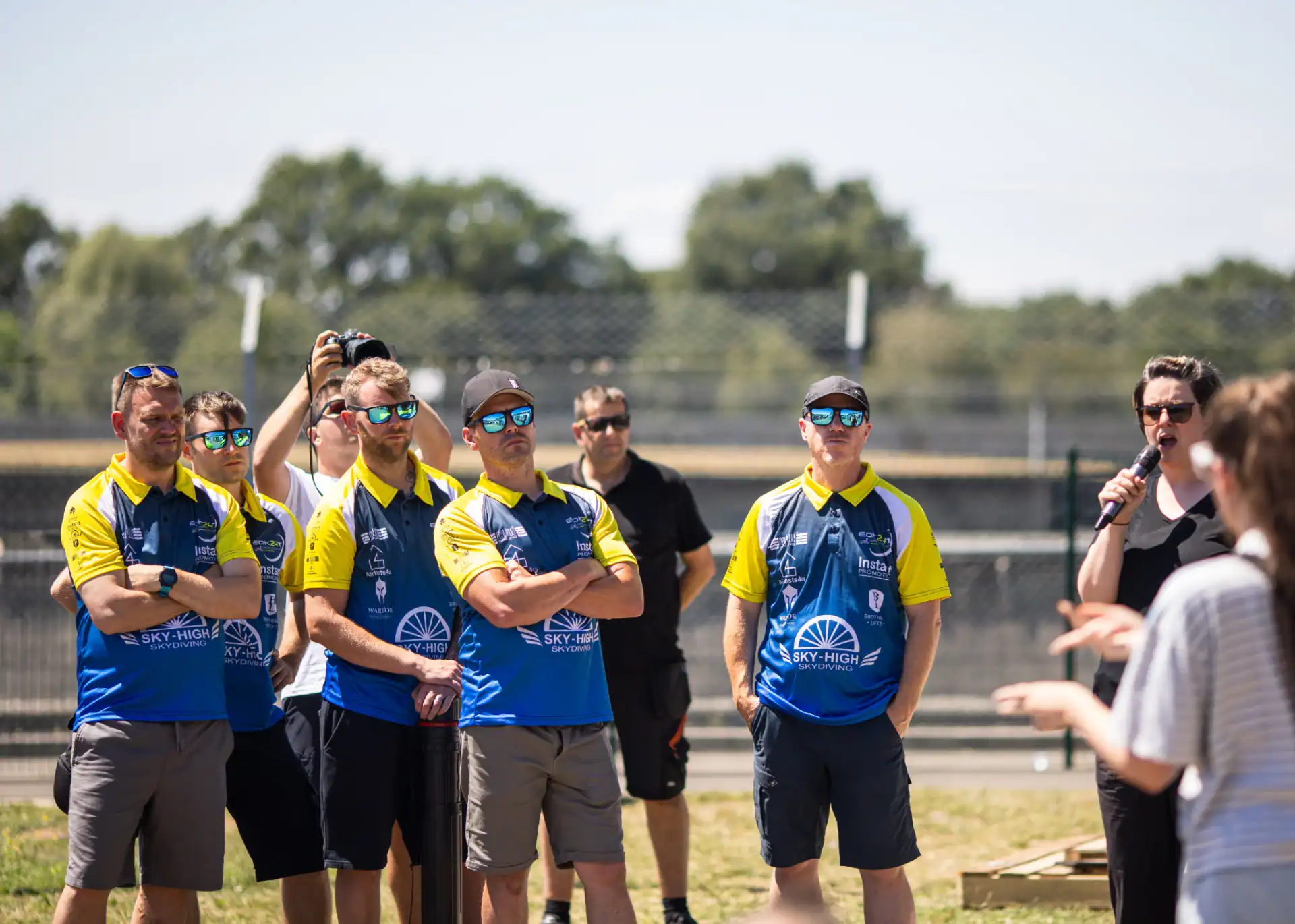 A group of people, some in matching blue and yellow shirts, stand outdoors listening to a person speak with a microphone. Trees and a fence are visible in the background.