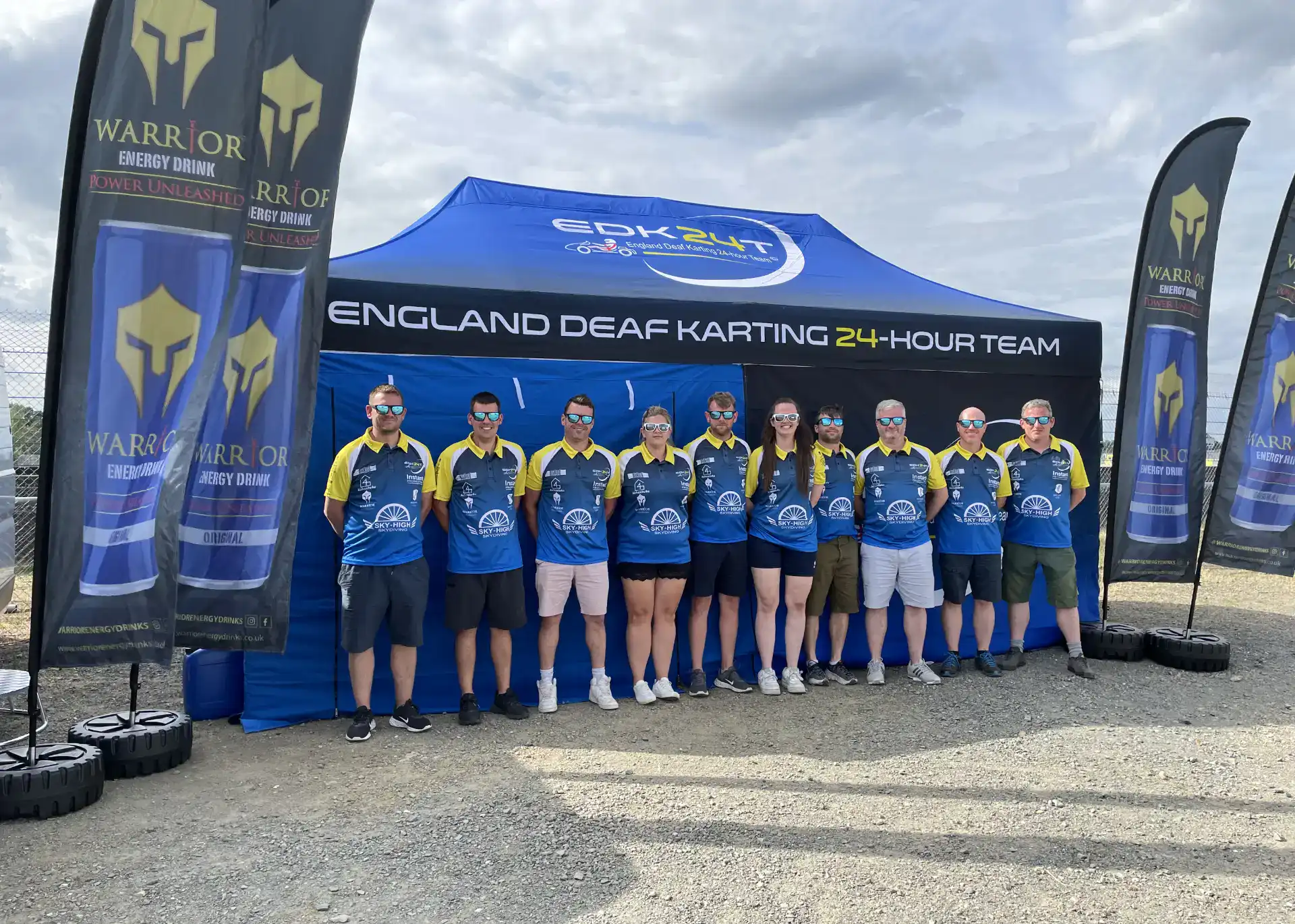 Ten people in matching blue and yellow shirts stand in front of a tent labelled "England Deaf Karting 24-Hour Team" with branded flags and tyres nearby.