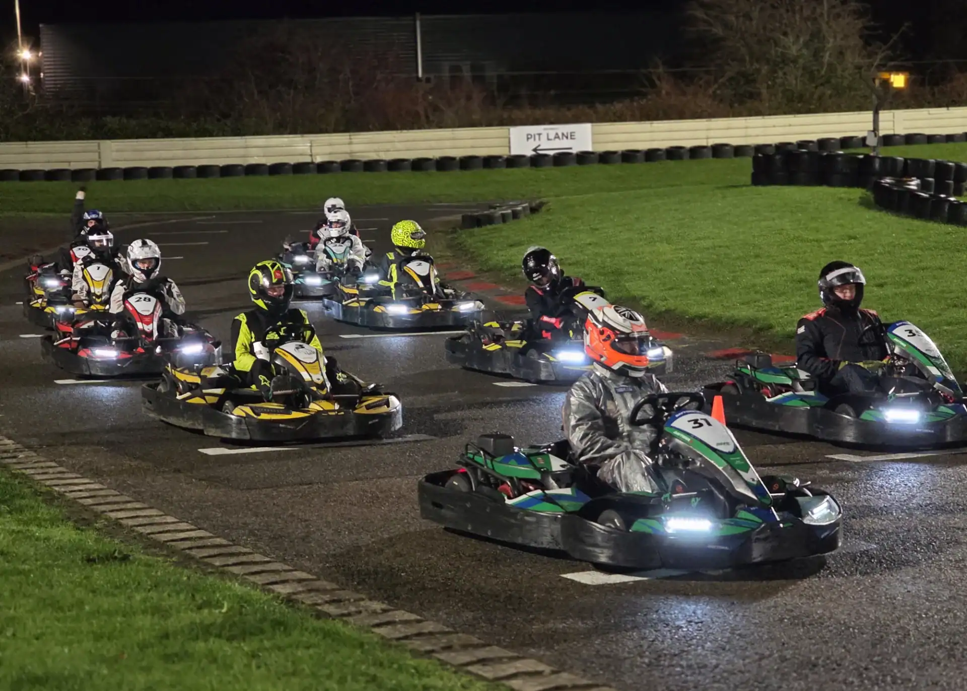 A group of people wearing helmets and racing suits drive go-karts on an outdoor track at night under artificial lighting.