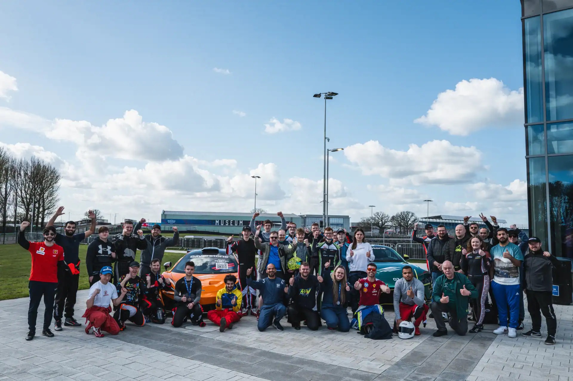 A large group of people pose and smile together outside near a sports car, with a building and trees in the background on a partly cloudy day.