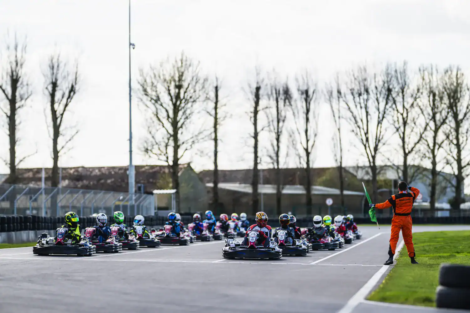 A group of go-kart racers lined up on a track as a race official in an orange suit waves a green flag to signal the start of the race.