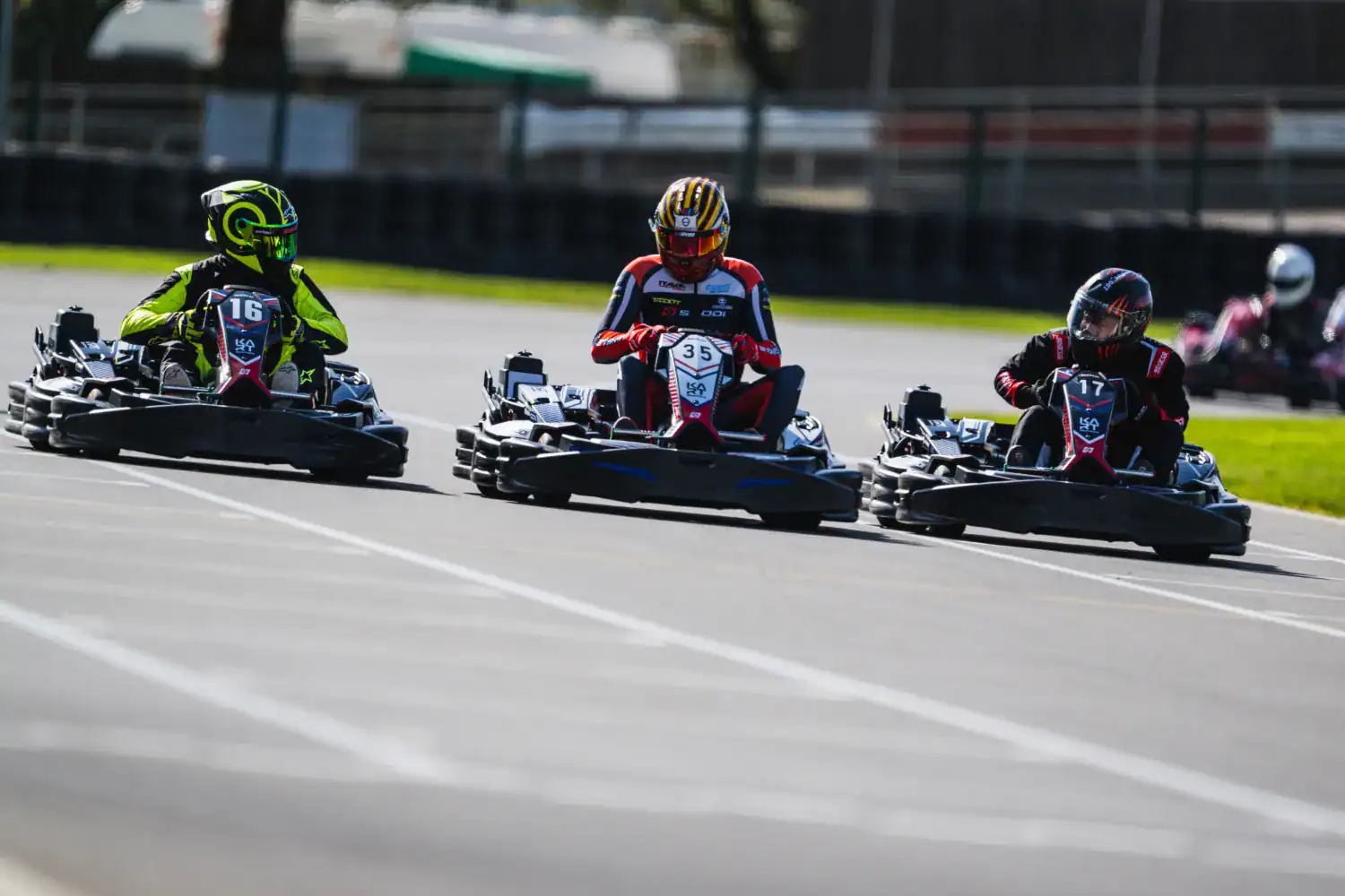Three people in racing helmets and gear drive go-karts side by side on an outdoor track during a race.