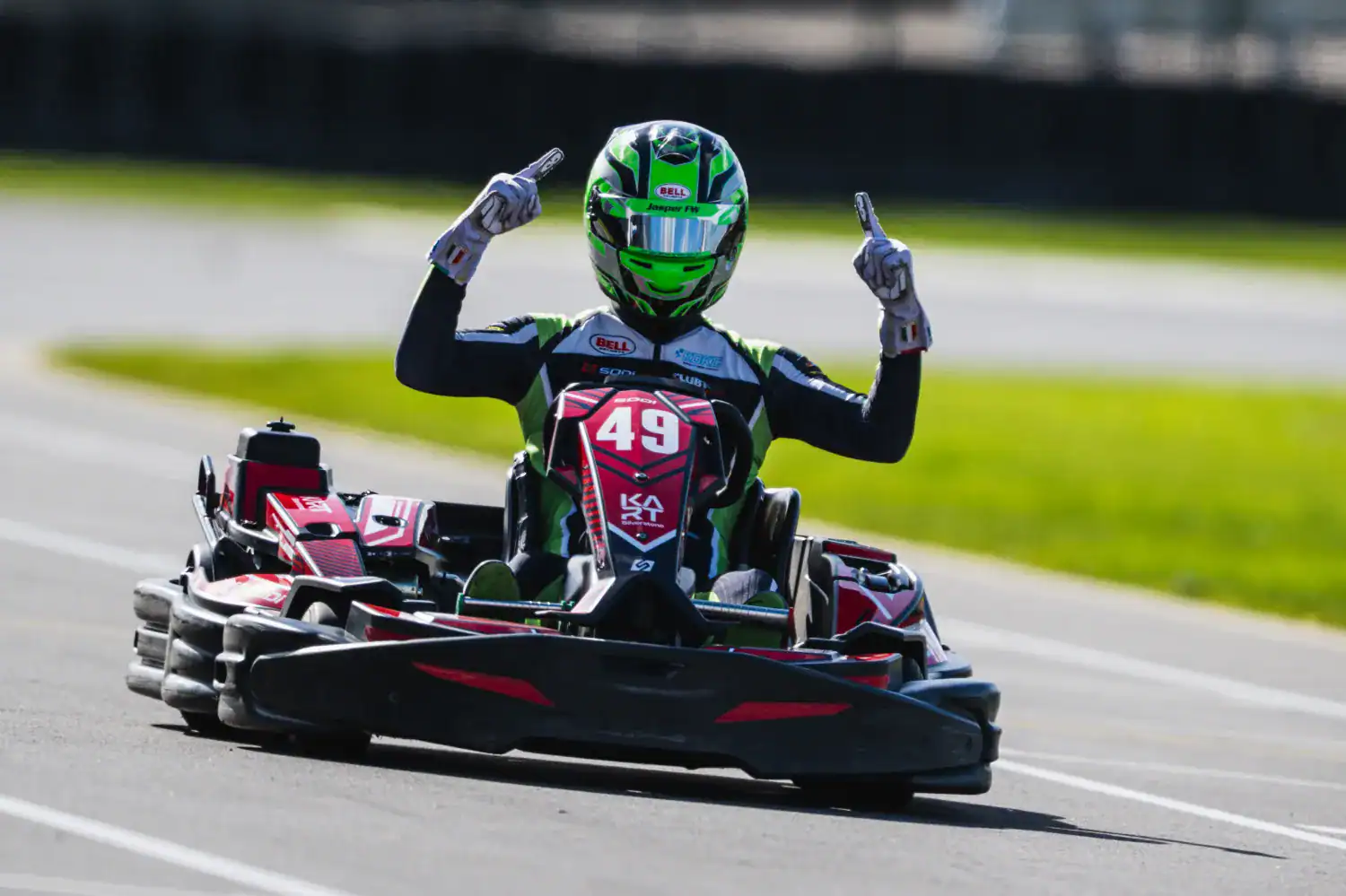 A go-kart driver wearing a green helmet and racing suit raises both hands with index fingers up whilst driving on a racetrack.