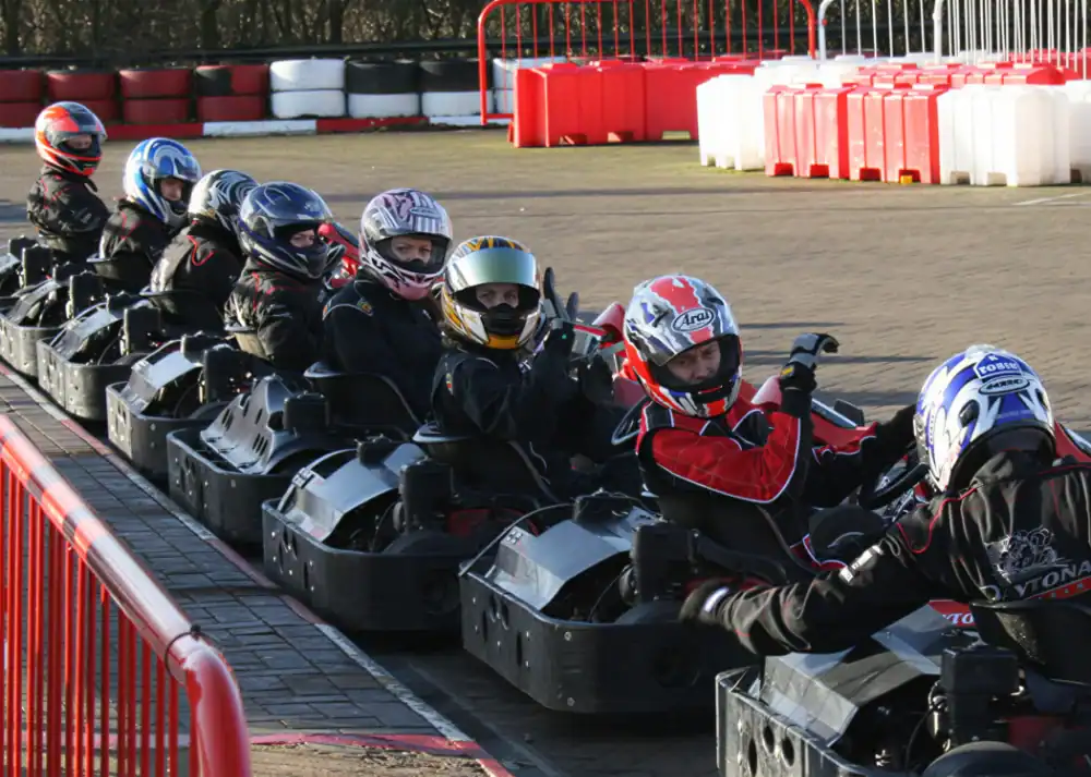 A group of people in helmets and racing suits sit in go-karts lined up on an outdoor track, ready to start a race.