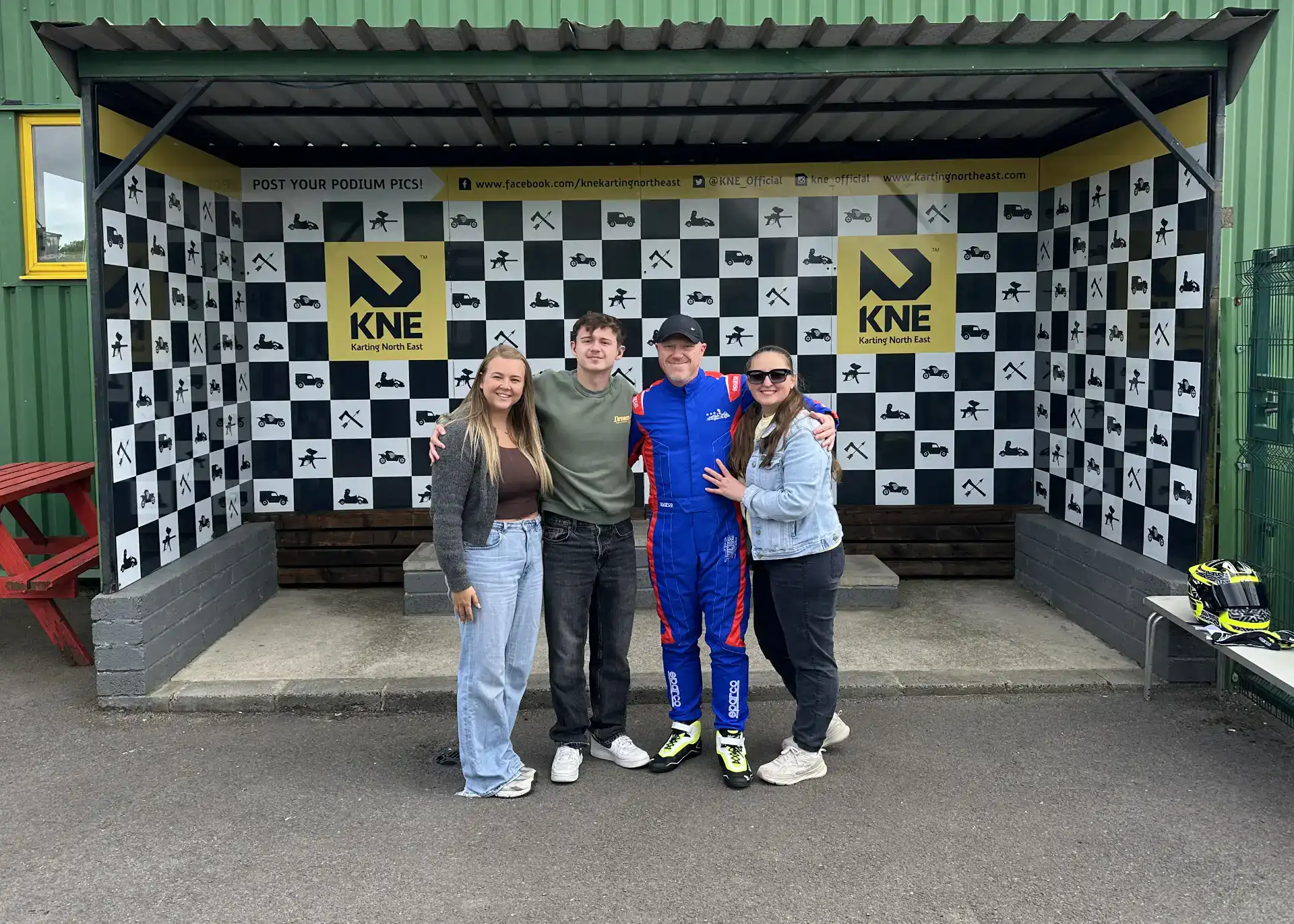 Four people stand together in front of a chequered podium backdrop at KNE karting, with one person wearing a blue racing suit and helmet nearby.