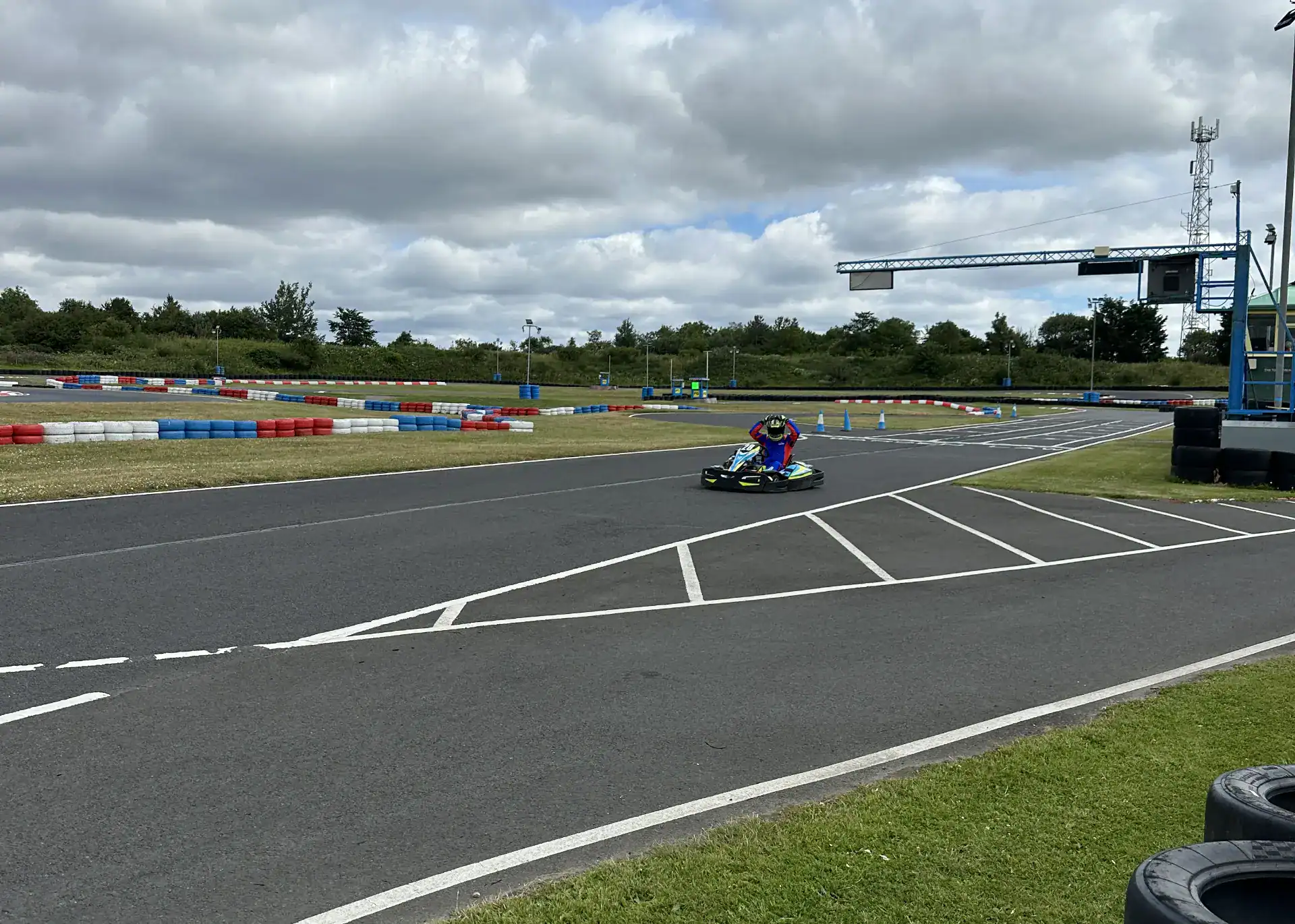 A person in racing gear drives a go-kart on an outdoor track with coloured barriers under a cloudy sky.