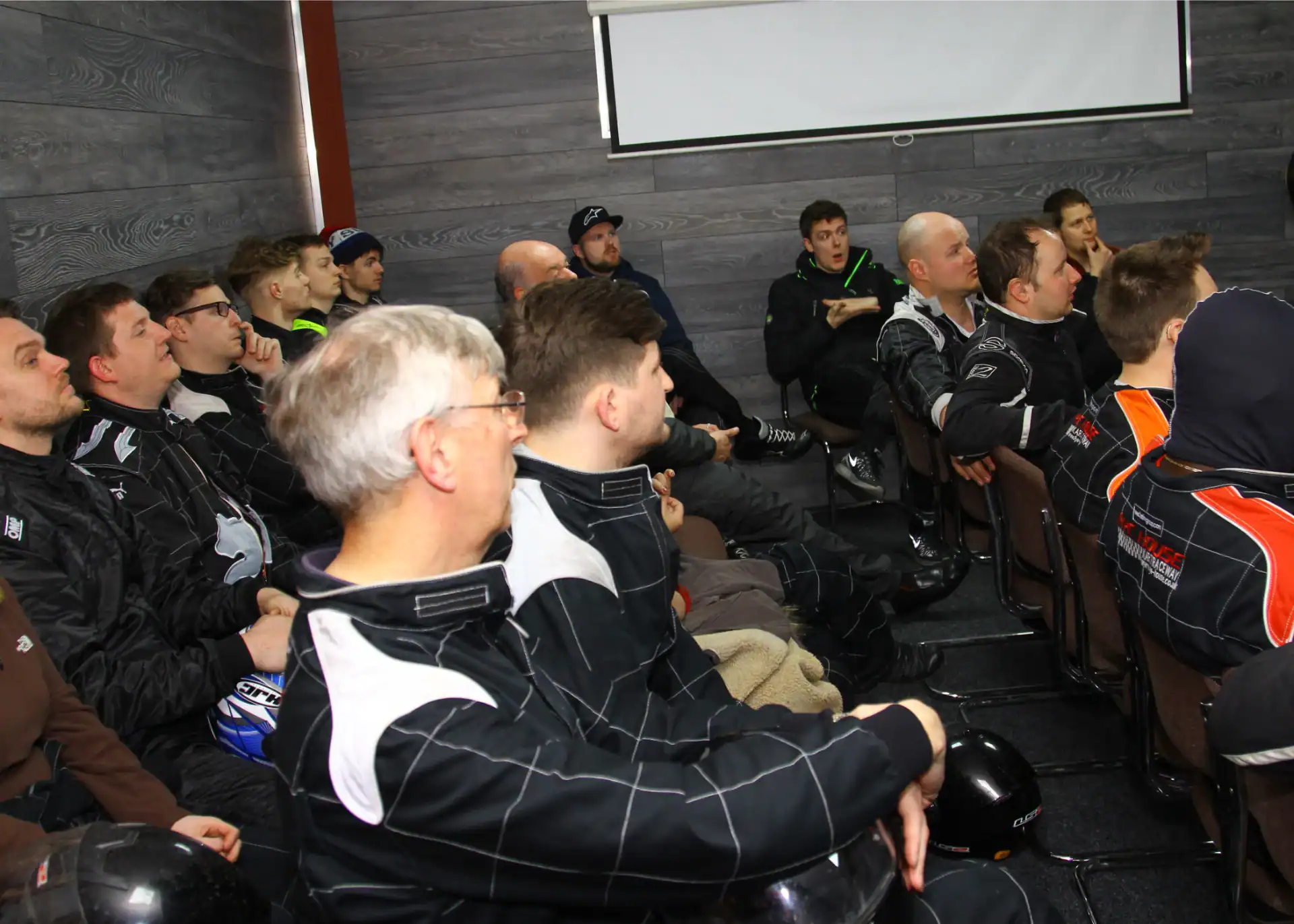 A group of people wearing racing suits sit in a room, attentively listening to someone out of frame, with helmets on their laps or nearby.