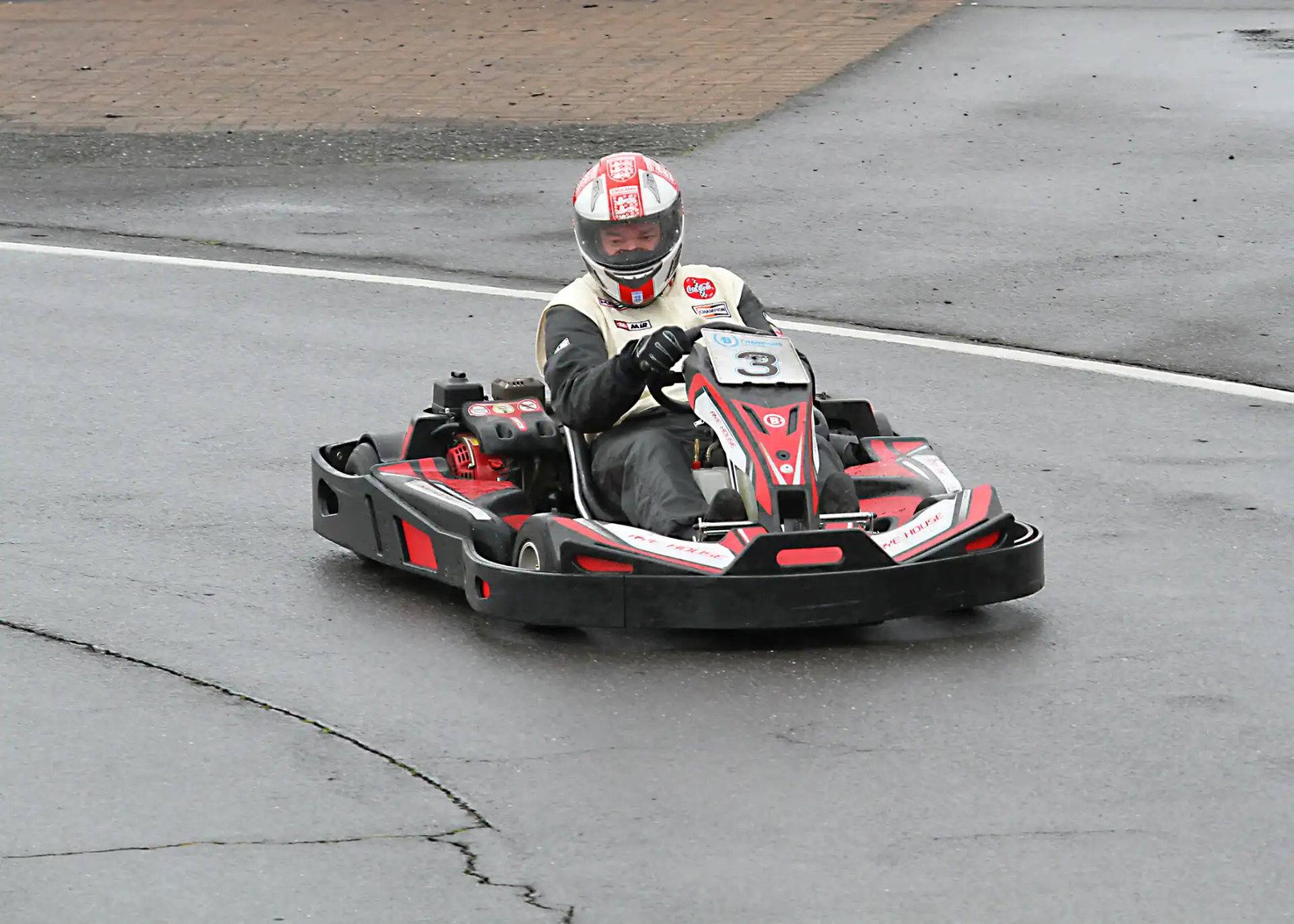 A person wearing a helmet and racing suit drives a black and red go-kart numbered 3 on a wet track.