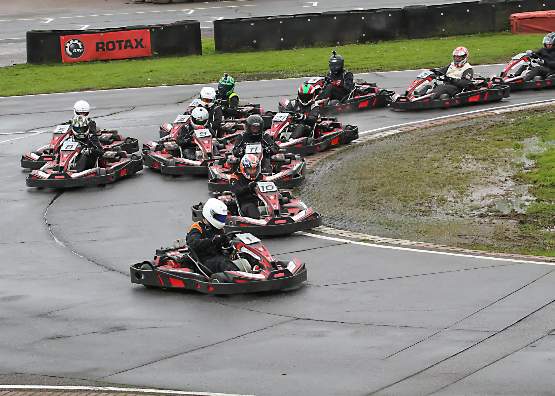 A group of people wearing helmets drive go-karts around a wet outdoor track, closely packed together as they navigate a bend.