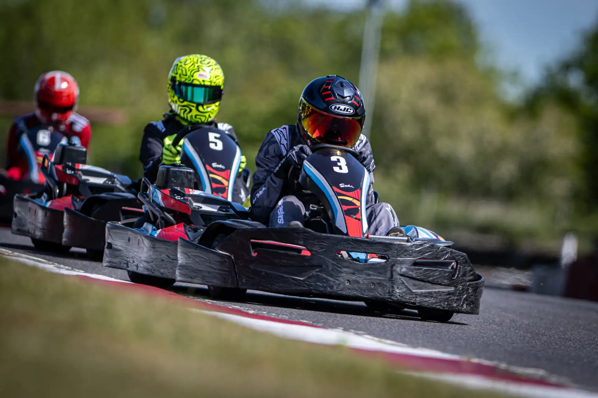 Three go-kart drivers race on an outdoor track, with driver number 3 in the lead and numbers 5 and 7 close behind. The background is blurred with greenery.