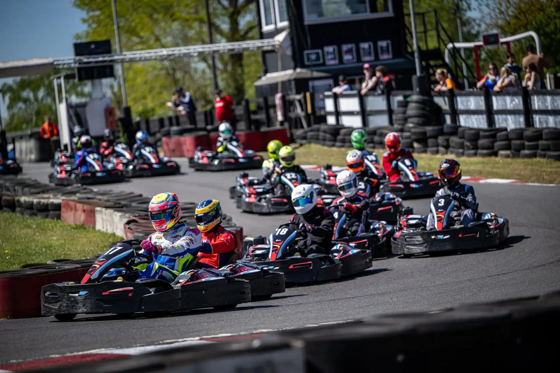 A group of go-kart racers in helmets navigate a bend on an outdoor track bordered by tyres, with spectators and trees in the background.
