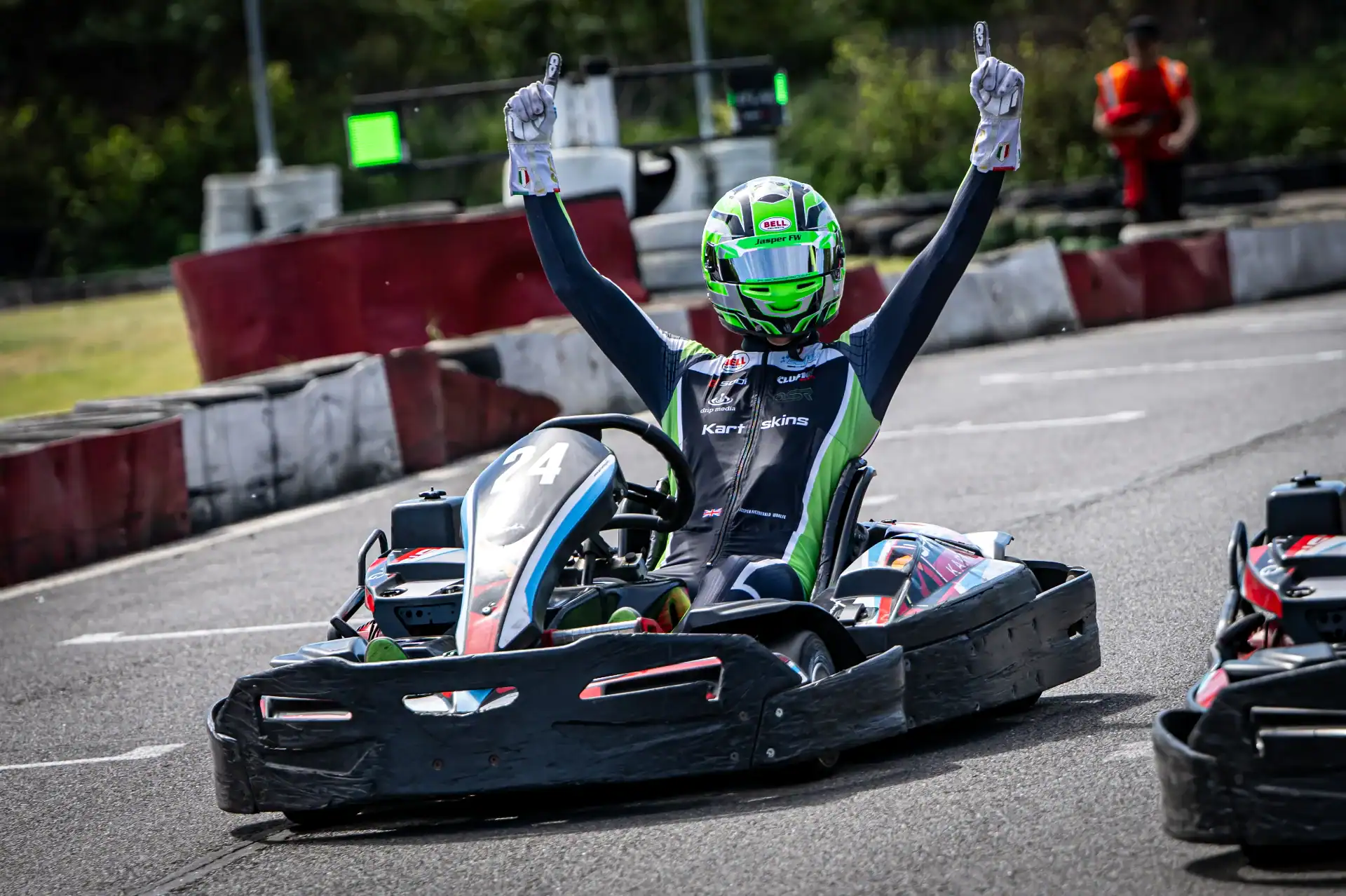 A go-kart racer wearing a helmet and racing suit raises both arms in celebration whilst crossing the finishing line on an outdoor track.