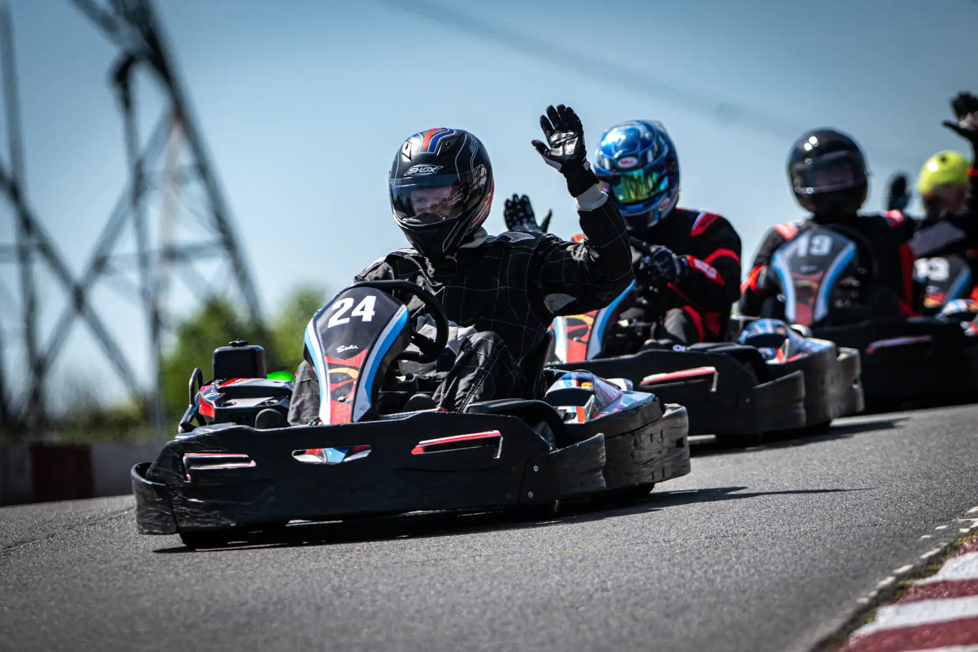 A go-kart racer wearing a black suit and helmet, numbered 24, waves whilst driving on a race track, followed by other racers.