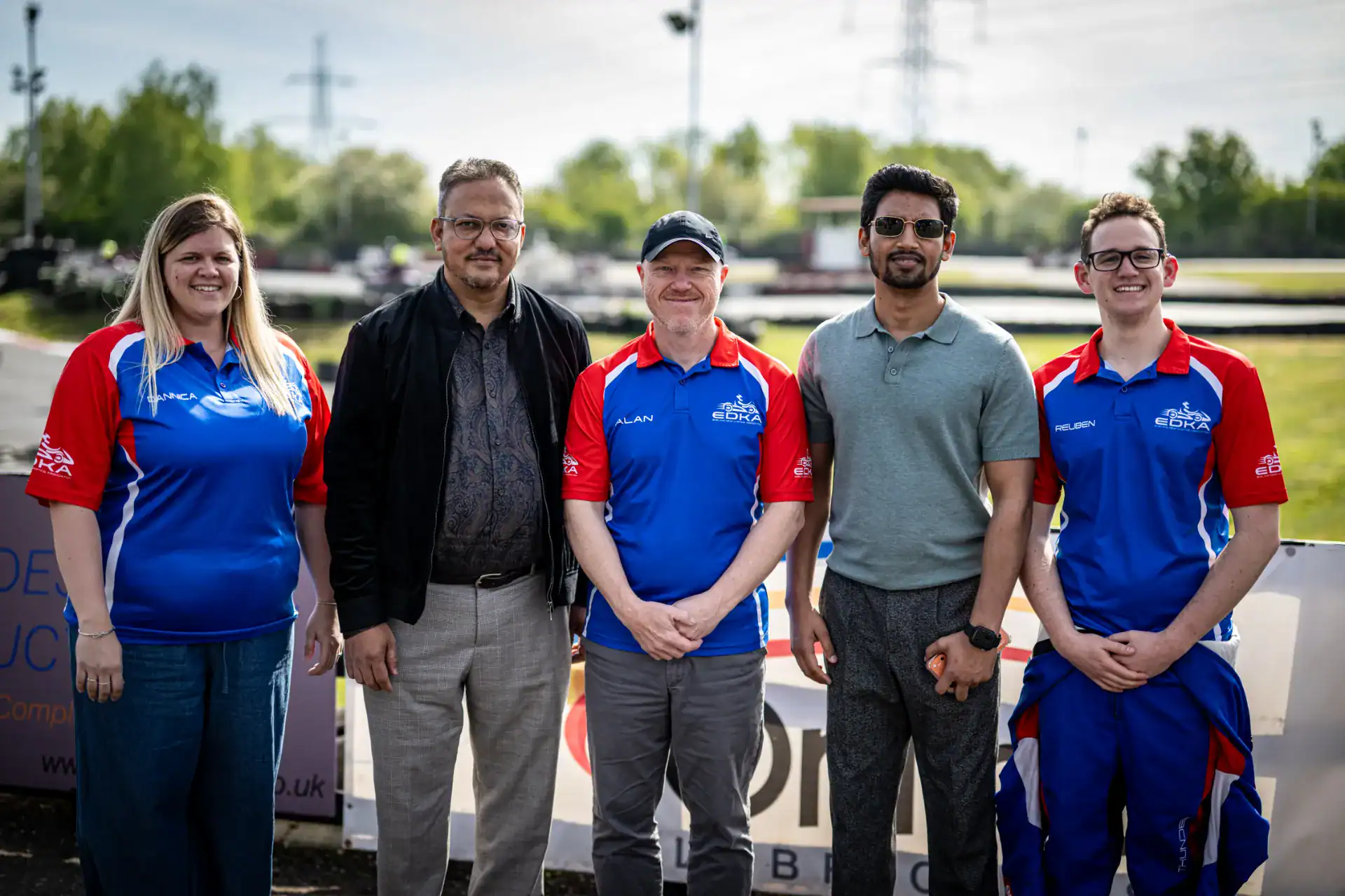 Five people stand outdoors, two in casual clothes and three in matching blue and red team shirts, posing for a group photo at a racetrack on a sunny day.