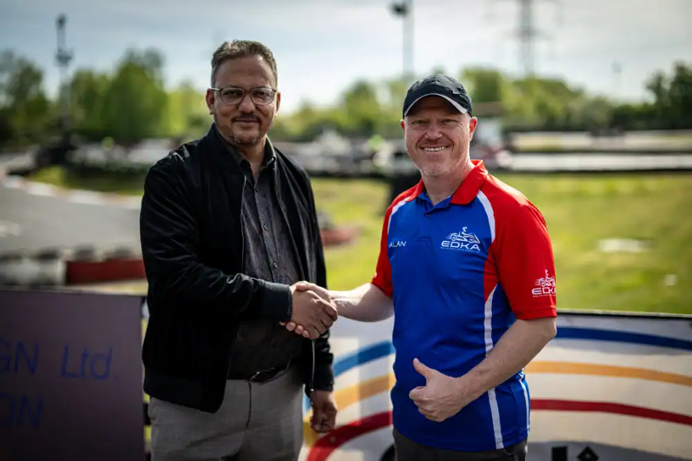 Two men shaking hands outdoors at a go-kart track, one in casual attire and the other in a blue and red shirt with a cap, both smiling at the camera.