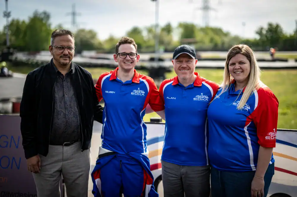 Four people stand together outdoors; three wear matching blue and red team shirts, and one is dressed in a dark jacket and shirt. A racetrack and greenery are visible in the background.