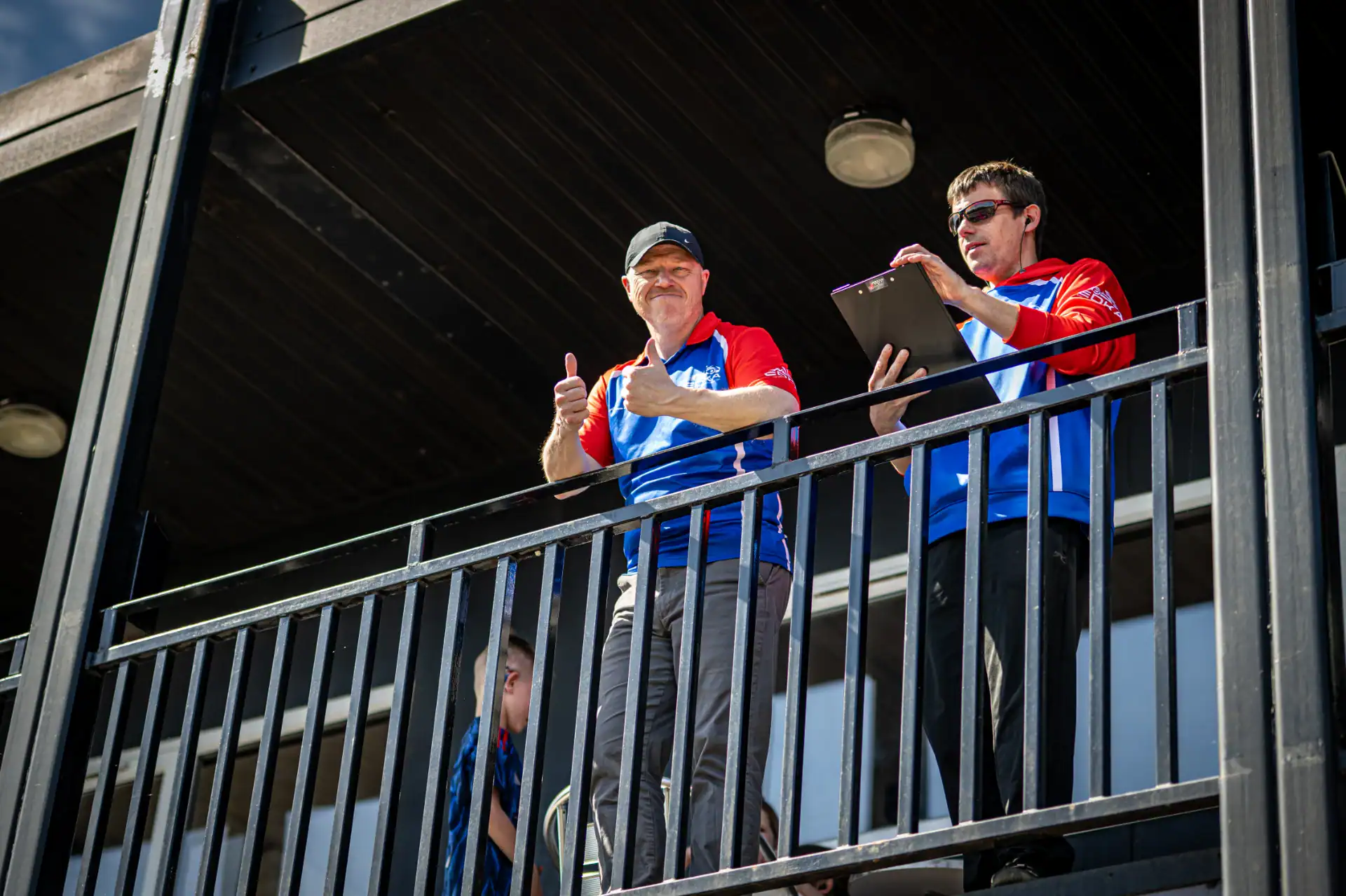 Two men in blue and red shirts stand on a balcony; one gives a thumbs-up whilst the other holds a clipboard.