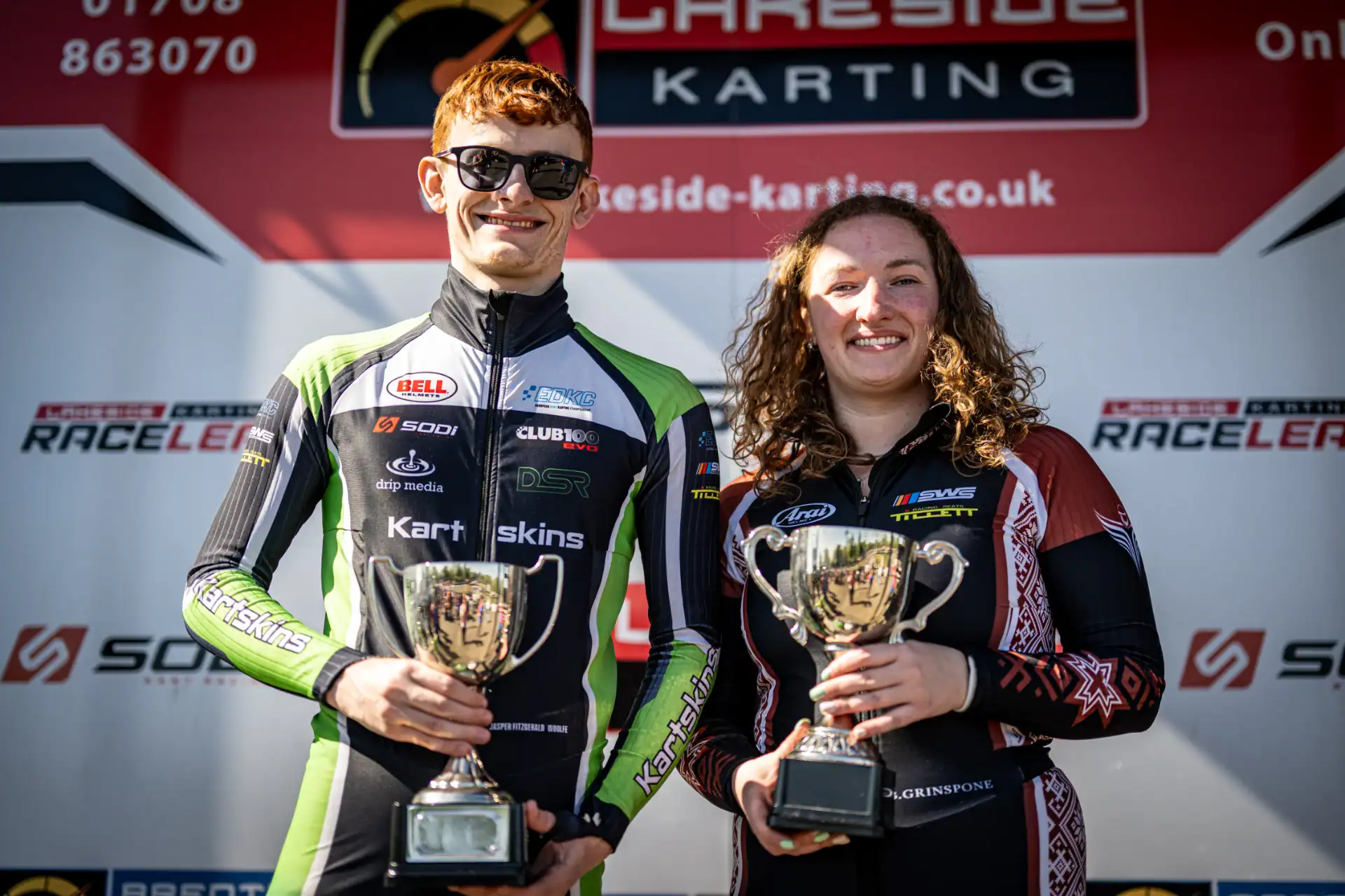 Two go-kart racers in racing suits stand side by side, smiling and holding trophies in front of a Lakeside Karting backdrop.