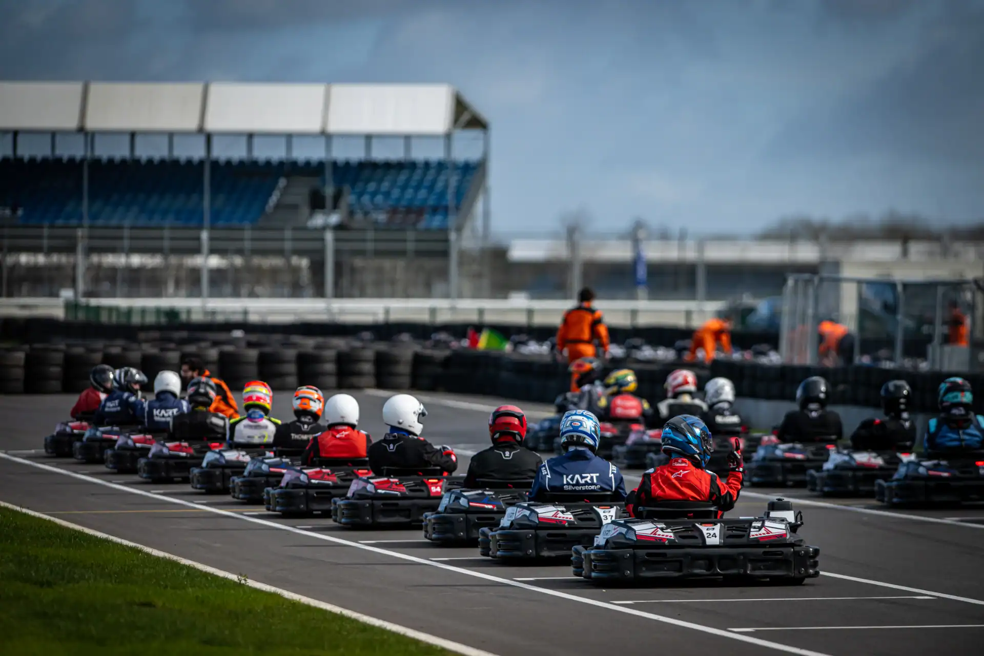 A group of go-kart drivers in helmets queue up on a race track, preparing for a race, with empty grandstands and track officials visible in the background.