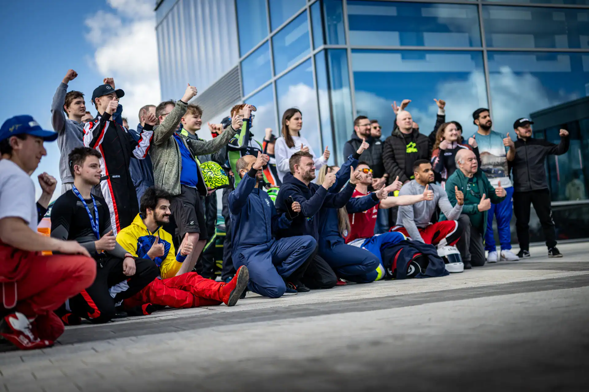 A group of people, some kneeling and others standing, pose together outside a modern glass building, many with raised fists or thumbs up gestures.