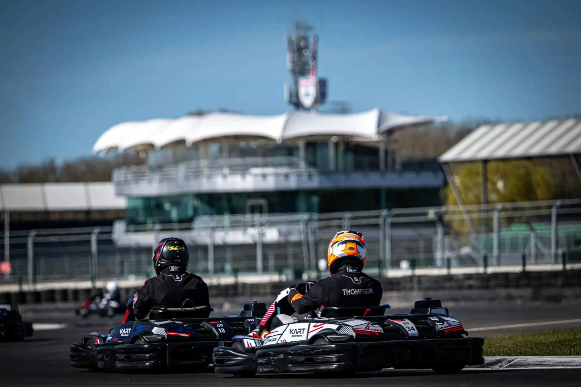 Two people in racing suits and helmets drive go-karts on an outdoor track with a modern building and fencing in the background.