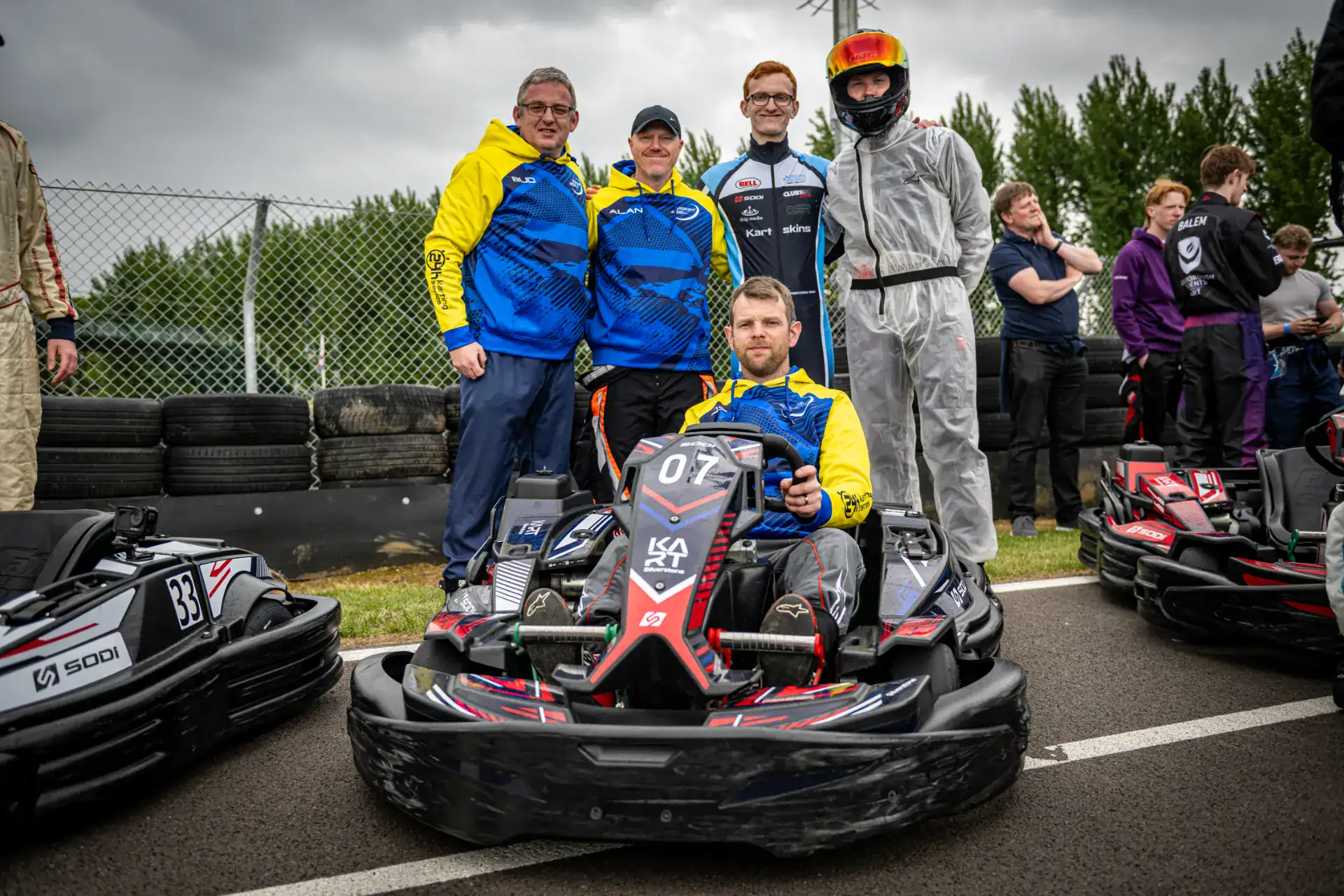 A group of five people, some in racing suits, stand and kneel behind a go-kart on a race track, with tyres and a chain-link fence in the background.