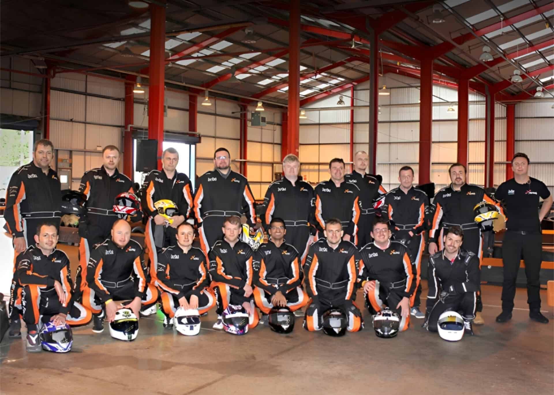 A group of 18 people in matching black and orange racing suits pose indoors with helmets, some standing and some kneeling in front.