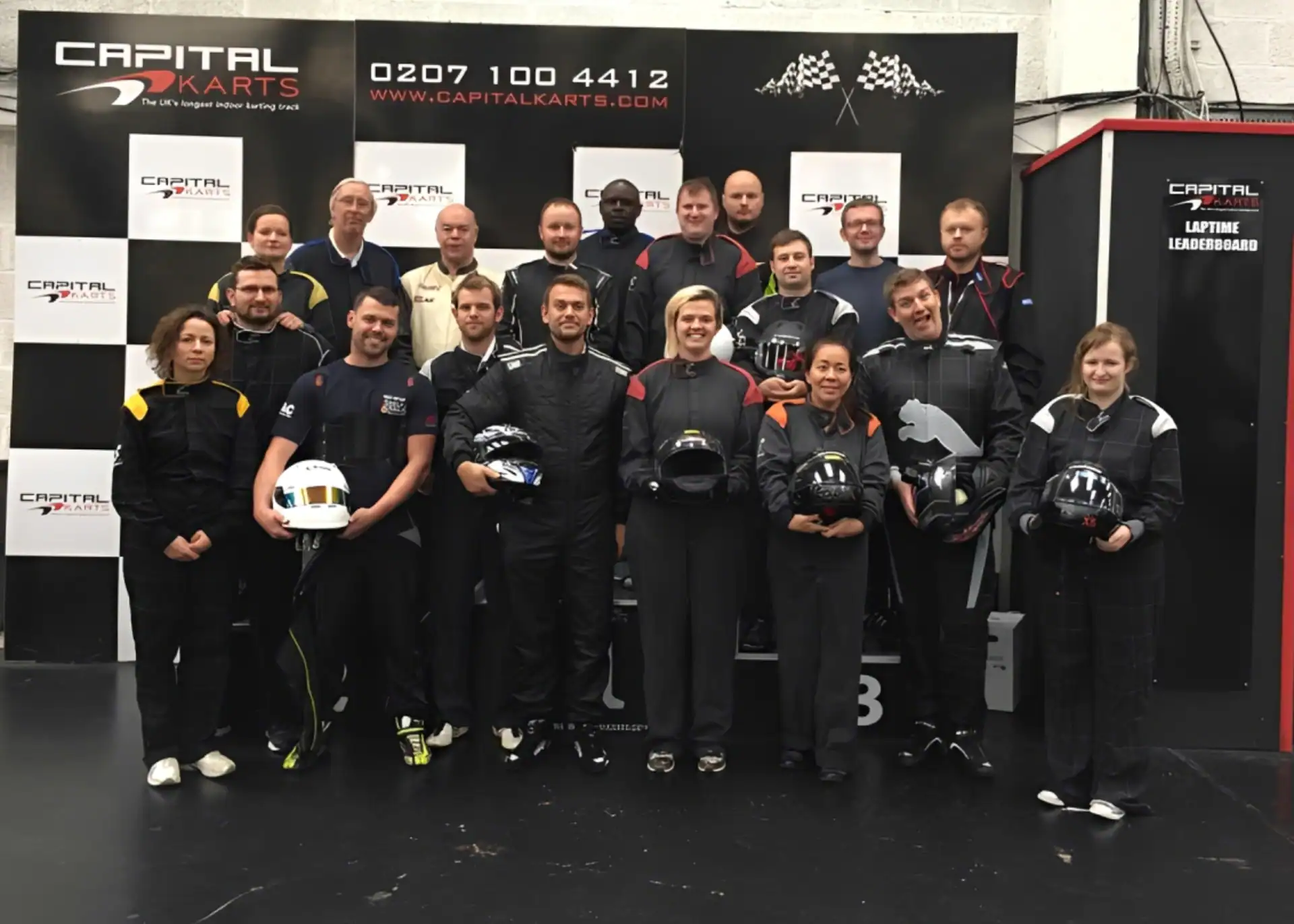 A group of people in racing suits pose with helmets in front of a Capital Karts backdrop, standing on an indoor go-kart track.
