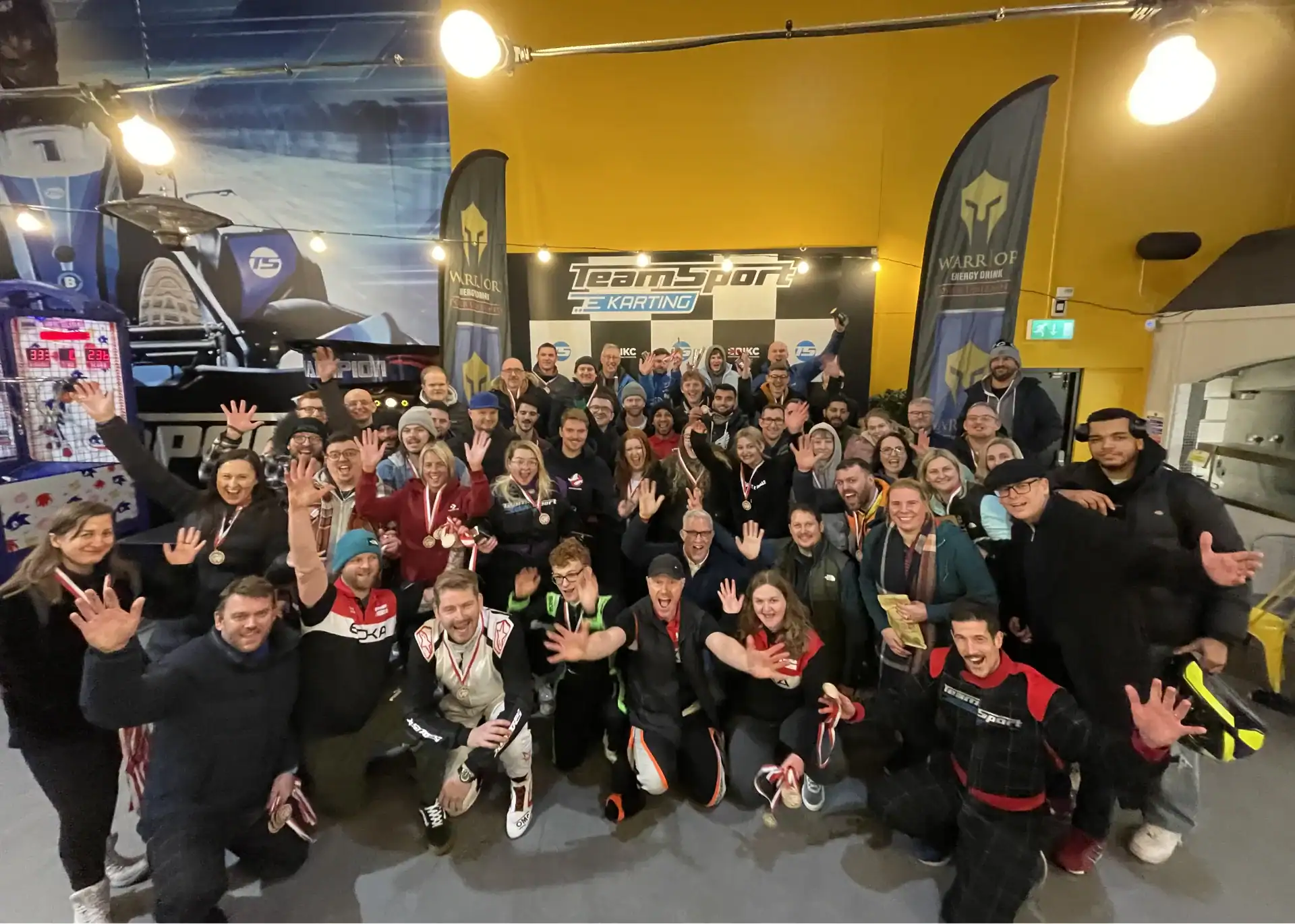 A large group of people pose together indoors at a go-karting venue, many smiling and waving at the camera, some wearing racing suits and medals.